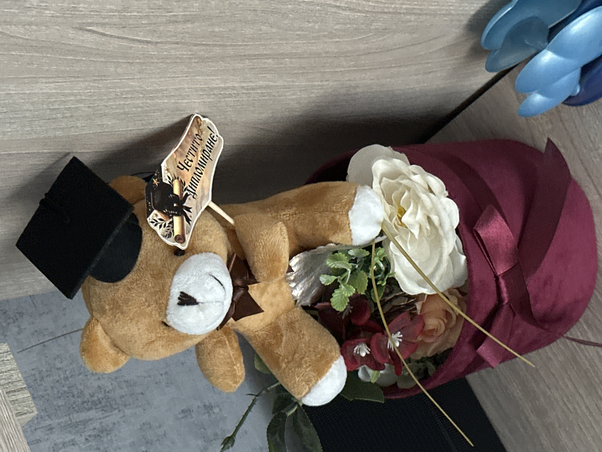 The image shows a teddy bear wearing a graduation cap, lying on a surface, possibly a table. The bear is part of a floral arrangement wrapped in burgundy fabric tied with a ribbon. The arrangement includes white and other colored flowers, along with greenery. A small sign that reads "Честито дипломиране!" (Congratulations on graduation!) is attached to the bear. The background appears to be a wooden surface. There is no indication of the location or any evidence suggesting that the image was taken in Lovech, Bulgaria.