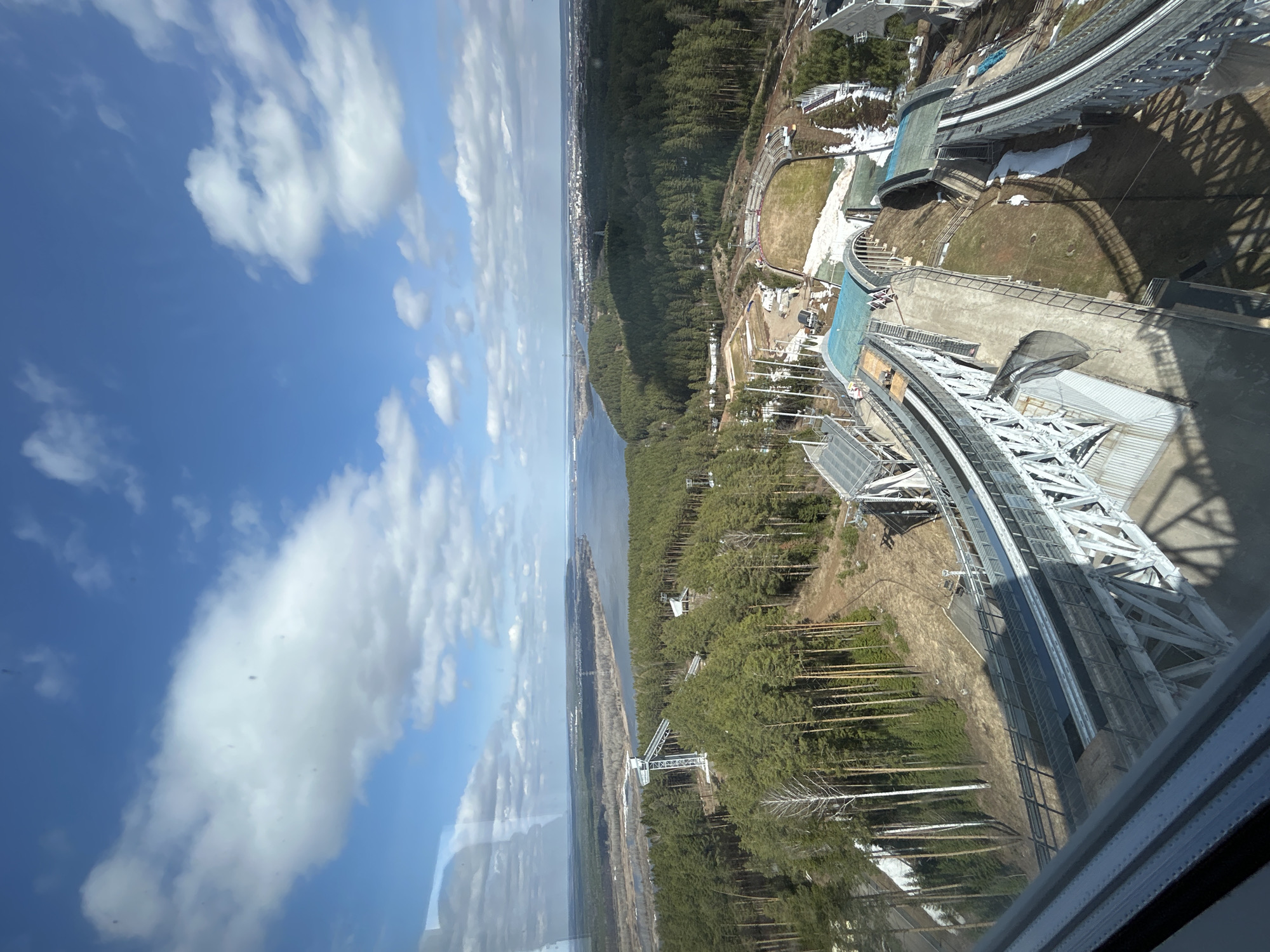 The image provides an elevated, panoramic view of a landscape featuring a large sports complex, dense coniferous forest, and a wide body of water, likely a river or lake, under a bright blue sky dotted with white clouds. The photograph appears to be taken from inside a building or platform, with a window frame and reflections visible in the foreground.
The prominent subject is a massive metal structure, resembling a ski jumping hill or a bobsled track, situated on the right side of the frame. This complex features a long, multi-level track supported by an intricate lattice framework, with associated smaller buildings and platforms. Patches of snow are still visible on the ground around the facility, suggesting a cool climate or early spring. The terrain immediately surrounding the complex consists of patchy grass and dirt, with some bare trees.
Beyond the sports infrastructure, a vast expanse of dark green pine forest covers the land, extending towards a wide, calm body of water that runs horizontally through the middle distance. On the far side of this water body, a distant town or city can be vaguely seen on the horizon. The sky is clear and bright, and the strong sunlight casts distinct shadows from the structures and trees. The overall impression is one of a large-scale outdoor sporting venue integrated into a natural, forested environment. The setting is daytime, with pleasant, sunny weather. No people or visible text are present in the image. The location is Прикамский, Russia.