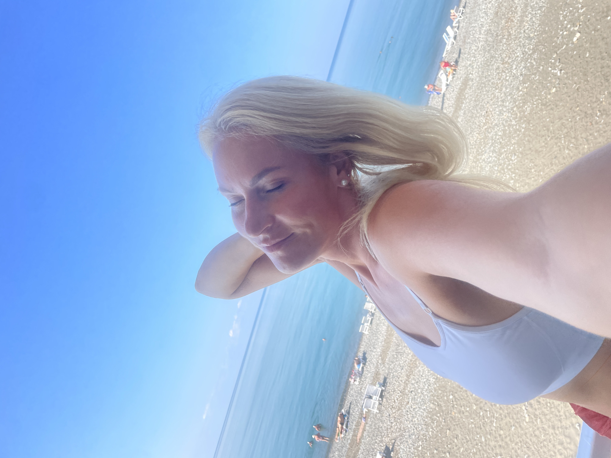 A blonde woman is captured in a selfie on a sunny day at a beach in Gagra, Abkhazia. She is wearing a white sports bra and has her eyes closed, with her right arm behind her head. The background shows a pebble beach with lounge chairs, people swimming and sunbathing in the clear blue sea, and a bright blue sky. The image is taken from a high angle, looking down at the beach.