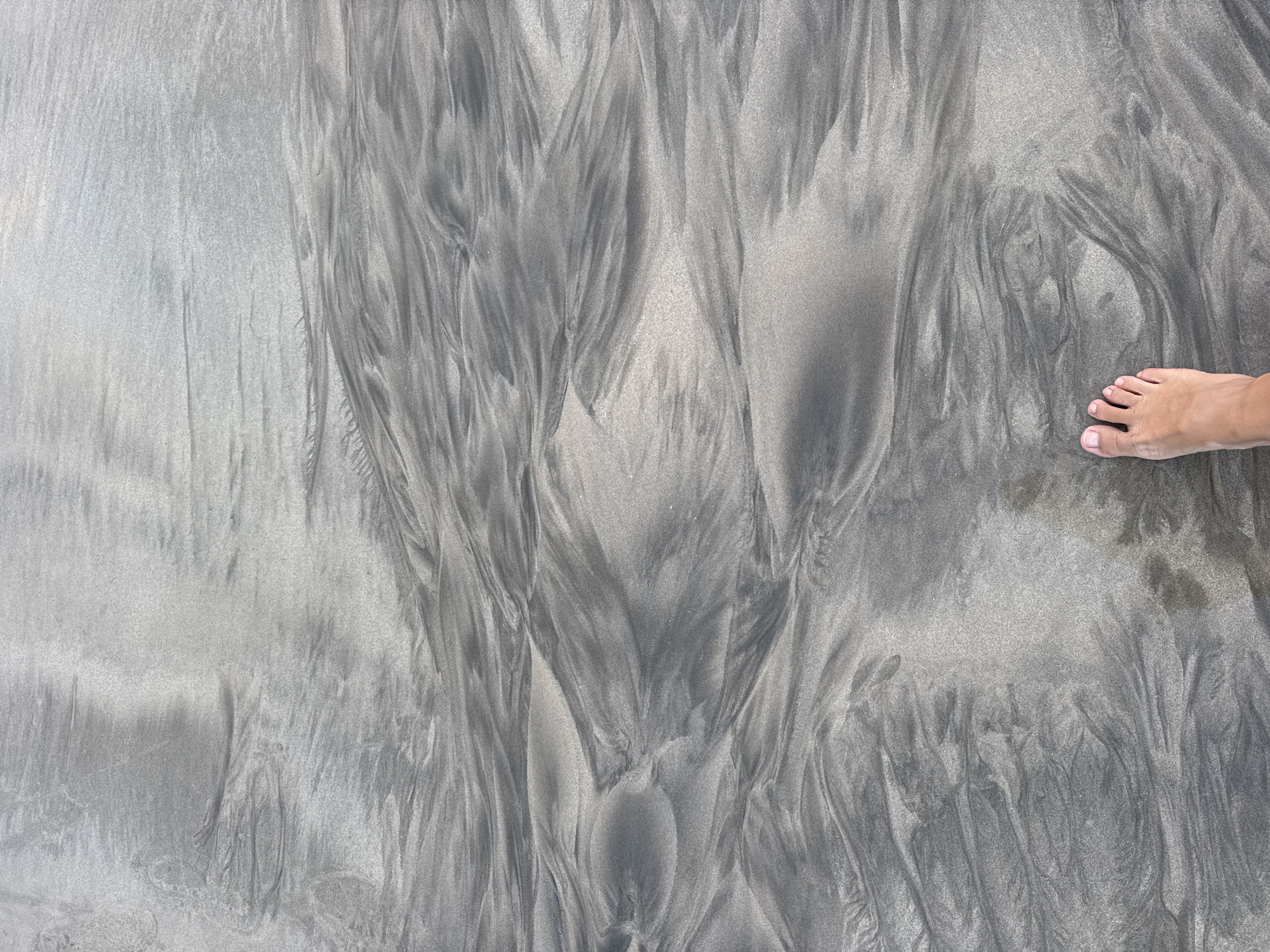 The image depicts a person's bare foot stepping onto a sandy beach. The sand displays intricate patterns, likely created by receding tides, with swirling lines of darker and lighter sand. The scene is an overhead shot of the beach, with the foot positioned in the lower right corner. The environment is natural, suggesting an outdoor setting, possibly Kuta Selatan, Indonesia, given the context. The overall impression is one of natural beauty and the vastness of the beach.