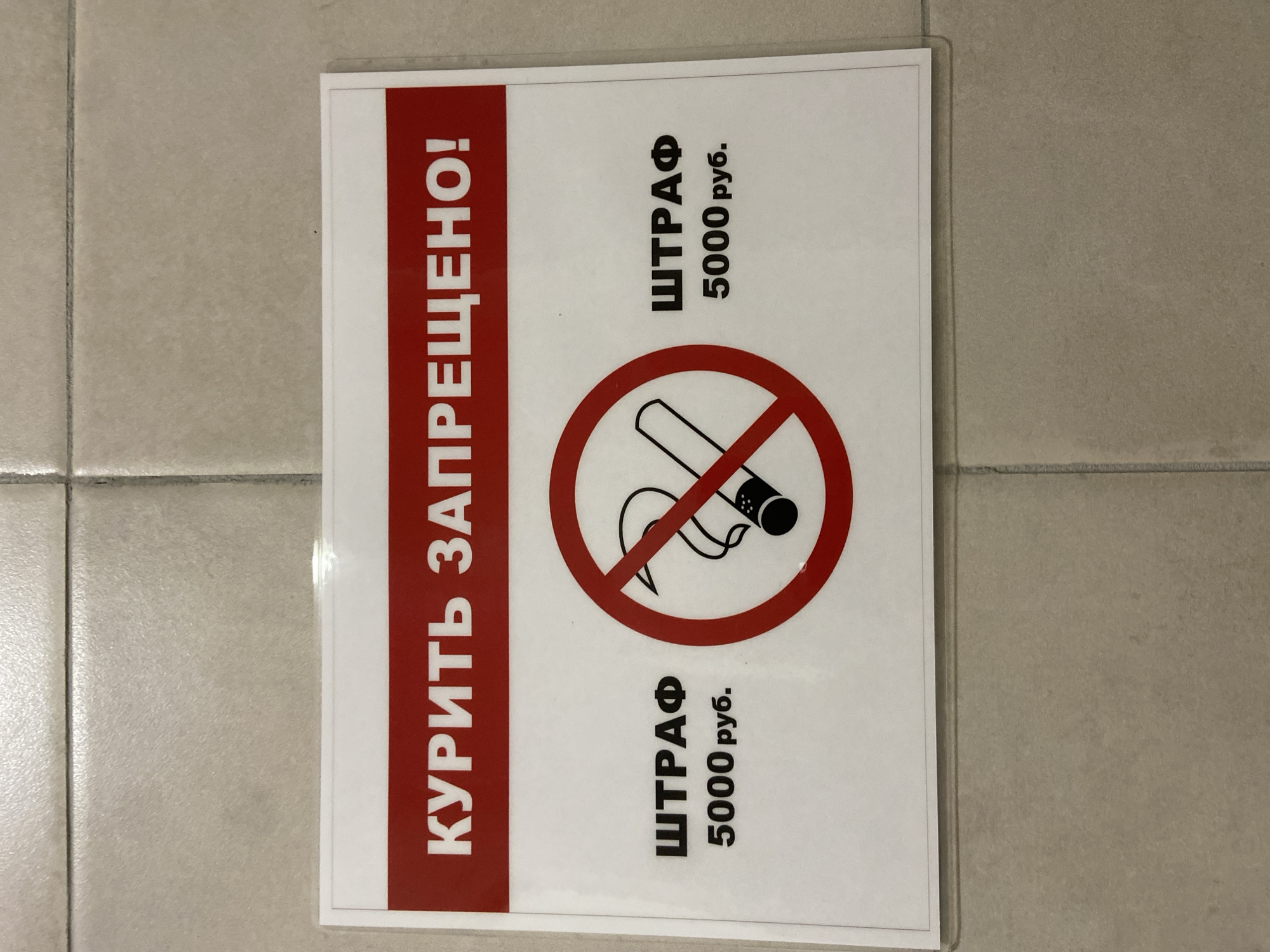 A sign on a tiled wall states "Smoking is prohibited!" in Russian text. Below the prohibition text, a red circle with a diagonal line through it encloses a stylized image of a cigarette with smoke. On the left and right sides of the prohibition symbol, text indicates a fine of "5000 rubles." The sign appears to be laminated and is attached to a wall constructed of large, light-colored square tiles, with visible grout lines. The lighting suggests an indoor environment. This sign is likely located in Yakutsk, Russia, given the Cyrillic text and the mention of rubles.