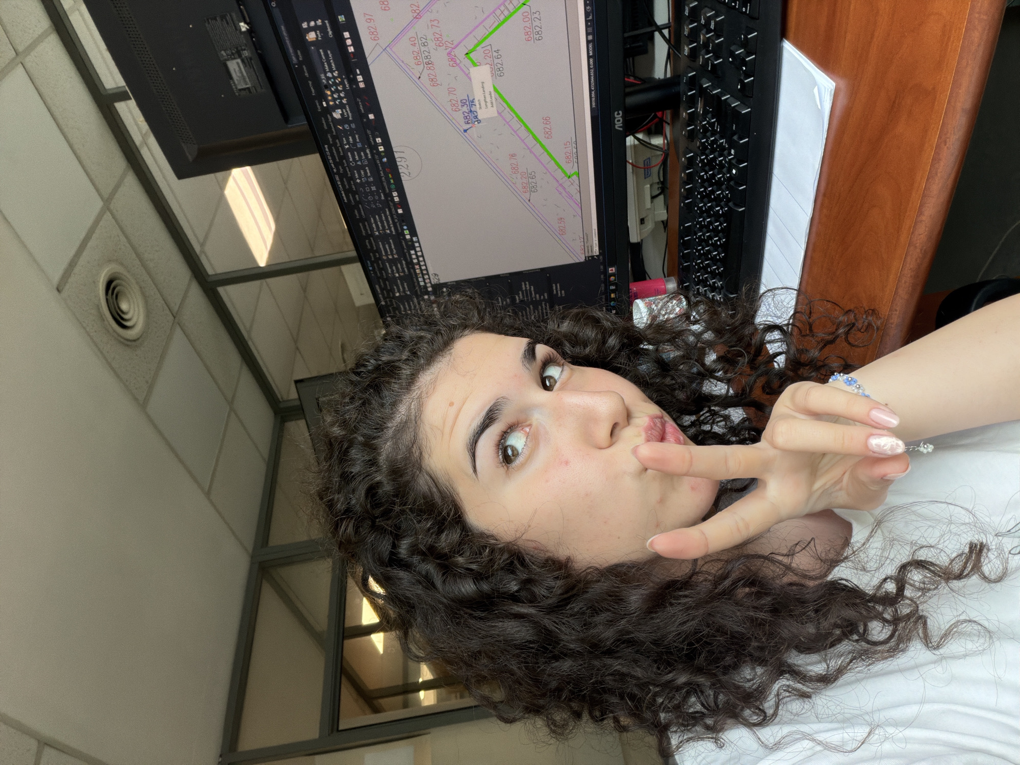 A young woman with curly brown hair is sitting at a desk in an office. She is looking towards the camera and holding up two fingers in a peace sign. She is wearing a white t-shirt and a bracelet on her left wrist. In front of her is a computer monitor displaying architectural plans. The monitor is on a desk made of wood. A keyboard is in front of the monitor. The room appears to be an office with fluorescent lighting and a drop ceiling. The overall environment suggests a professional workspace.