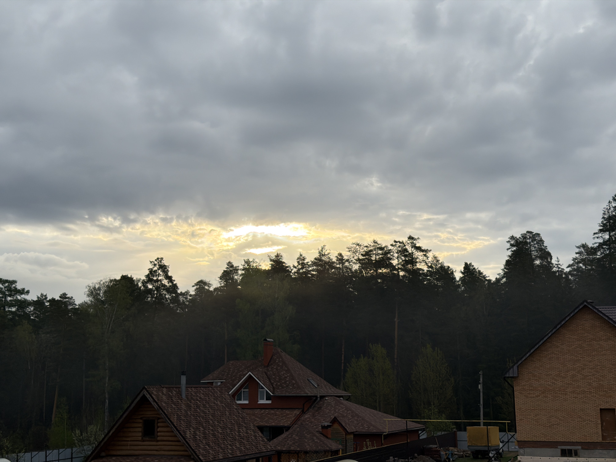 The image captures an overcast sky over what appears to be a residential area bordering a forest. The sky is filled with gray clouds, but there is a patch of lighter sky with a hint of yellow sunlight breaking through near the horizon. Below the sky and the band of trees is an assortment of rooftops, primarily consisting of houses. The houses exhibit varied architectural styles with different roofing materials and colors. The house on the right side of the image is constructed of brick and has a light brown color.
The environment is likely a suburb or residential area on the outskirts of a city, given the proximity to the forest. The presence of houses suggests a settled community, while the lighting conditions, with low sunlight and overcast skies, create a subdued atmosphere. The image was taken in the city of Chaikovsky, Russia.