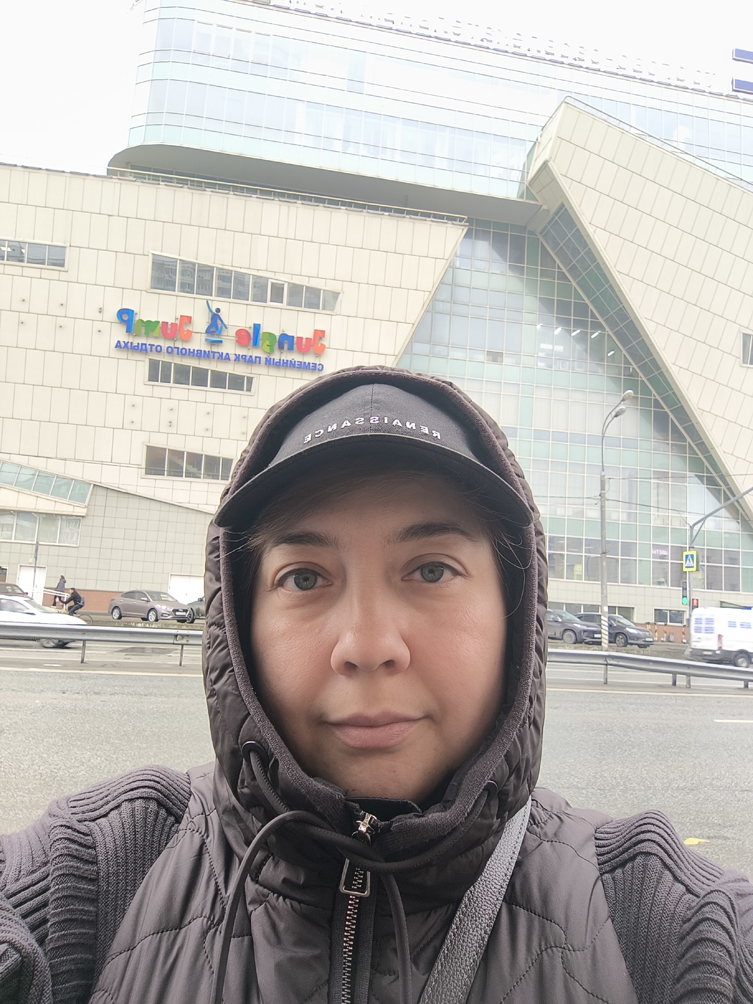 The image depicts a person taking a selfie in front of a building with a sign that reads "Jungle". The building has a modern architectural design with geometric shapes and glass panels. The person is wearing a black hooded jacket and a baseball cap with "RENAISSANCE" written on it. There is a road with vehicles in the background, and a pedestrian crossing sign is visible. The location appears to be in an urban environment.