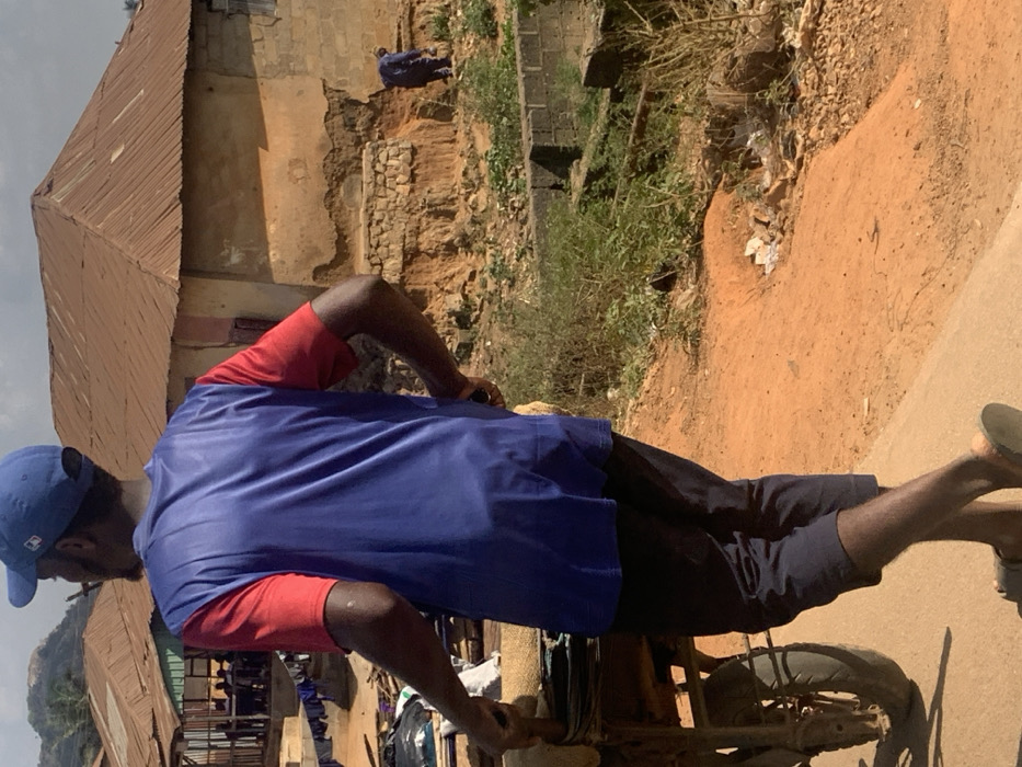 The image displays a daylight scene in Egbedi, Nigeria. In the foreground, a dark-skinned male, viewed from the back, walks left while pushing a two-wheeled cart. He wears a blue baseball cap, a blue t-shirt with red short sleeves, dark trousers, and light-colored sandals. The cart, equipped with a dark-colored wheel and tire, carries stacked items, possibly wrapped goods or documents.
The ground is an unpaved reddish-brown dirt path, with a visible concrete or paved edge to the immediate left. To the right of the path, the terrain drops off into an area of disturbed reddish soil and sparse green vegetation.
In the midground, a single-story building with light-brown, textured walls and a rusted, corrugated metal pyramidal roof stands on the right. Its foundation exhibits significant erosion, exposing layers of earth and stonework. A small, dark opening, possibly a window, is visible on its side. Further into this eroded area, a second, less distinct figure, dressed in blue, appears to be stooping or working. To the left, another single-story structure with a similar rusted roof is visible, with laundry hanging on a line nearby.
The background features distant, hazy hills or rock formations beneath a light blue sky with scattered white clouds.