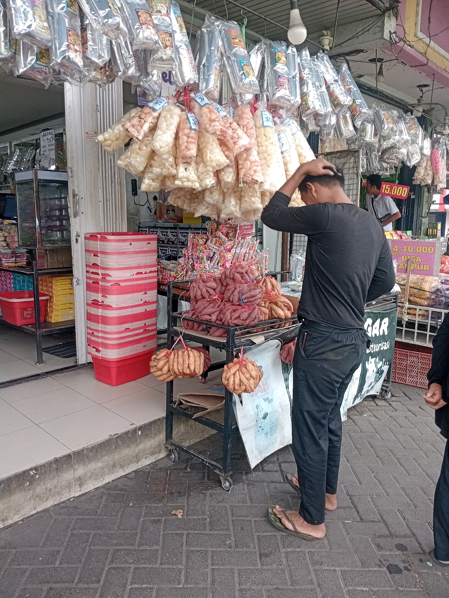 A man in a black long-sleeved shirt and black pants stands in front of a street stall in Lawang, Indonesia, with his hand on his head. The stall displays a variety of goods, including bags of snacks hanging from the ceiling, a cart with piles of sausages, and a display of colorful lollipops. A sign with "15.000" is visible. In the background, another man in a light-colored shirt is seen. The ground is paved with dark bricks. The overall scene depicts a bustling market environment.