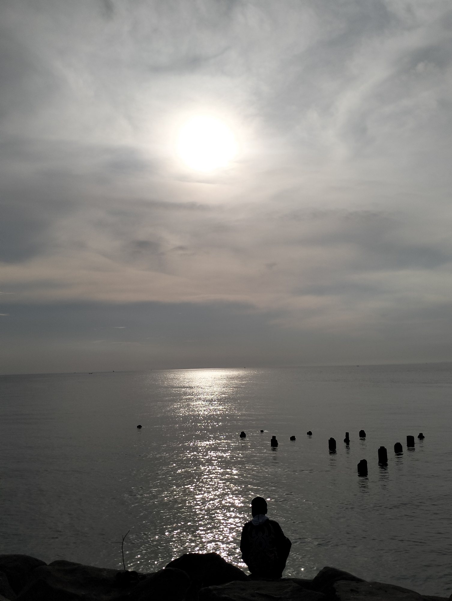 A wide-angle, outdoor shot depicts a body of water under a cloudy sky with a bright, diffused sun high in the frame. The water reflects sunlight, creating a shimmering effect across its surface. Several short, dark cylindrical posts are partially submerged in the water, spaced irregularly. In the foreground, a person is sitting with their back to the viewer on what appears to be a rocky embankment. The figure is silhouetted, obscuring details of clothing and features. The overall tone of the image is somber due to the muted colors and obscured light. There is no indication in the image to determine a specific location in Lhokseumawe, Indonesia.
