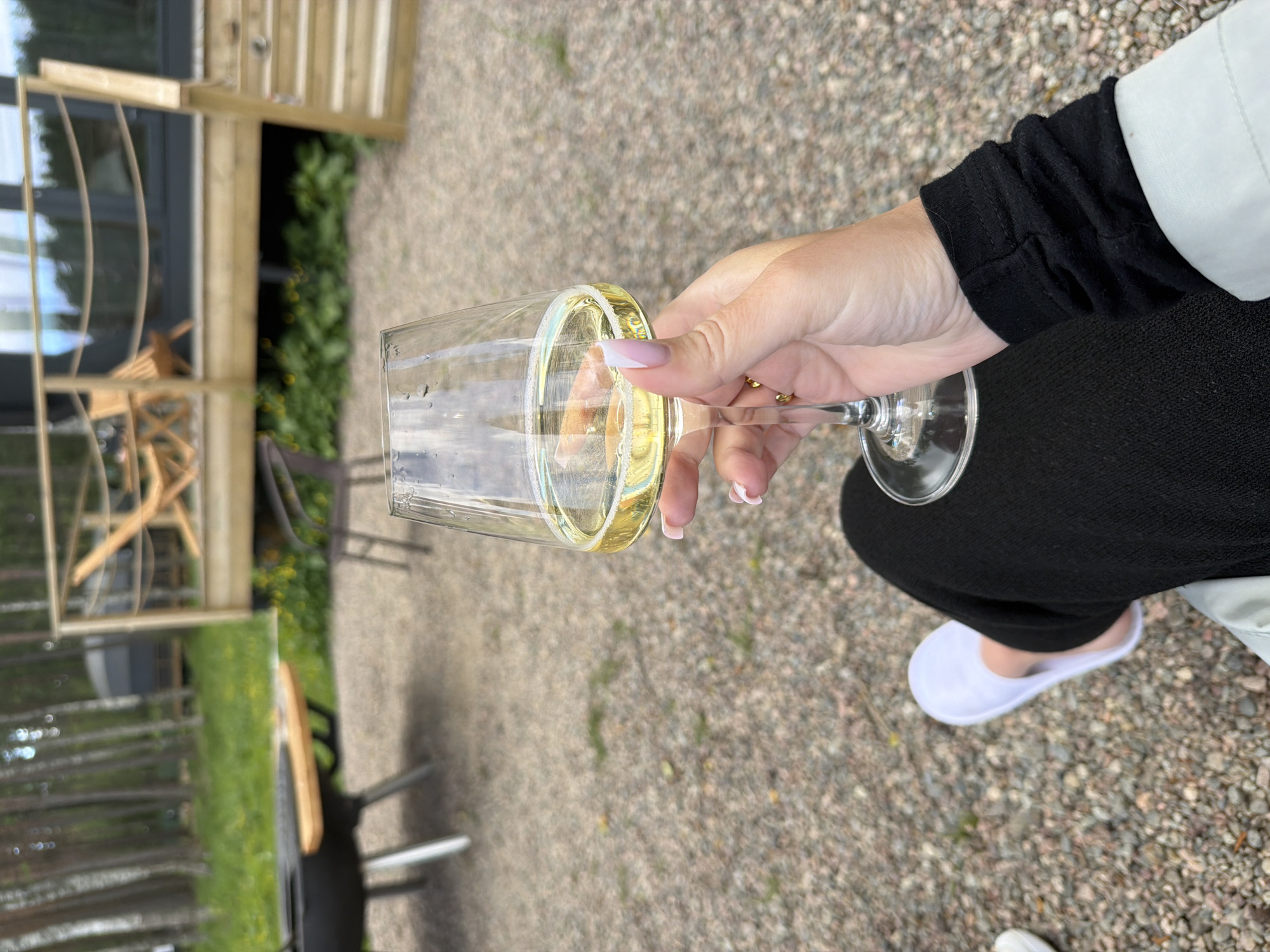 A person's hand holds a wine glass filled with a light-yellow liquid, suggesting white wine, outdoors. The hand has manicured nails with a French tip design and a small gold ring on the finger. The person is wearing black clothing and a white slipper. In the background, a gravel-covered area leads to a wooden deck of a building, possibly a cabin or guesthouse. A grill and table are visible to the left, and trees and vegetation form the backdrop, indicating a natural setting. The context of Solov'evo, Russia, suggests this scene is likely taking place in a rural or semi-rural area, perhaps a vacation spot or a private residence with outdoor amenities. The lighting and atmosphere suggest daytime, possibly late afternoon or early evening.