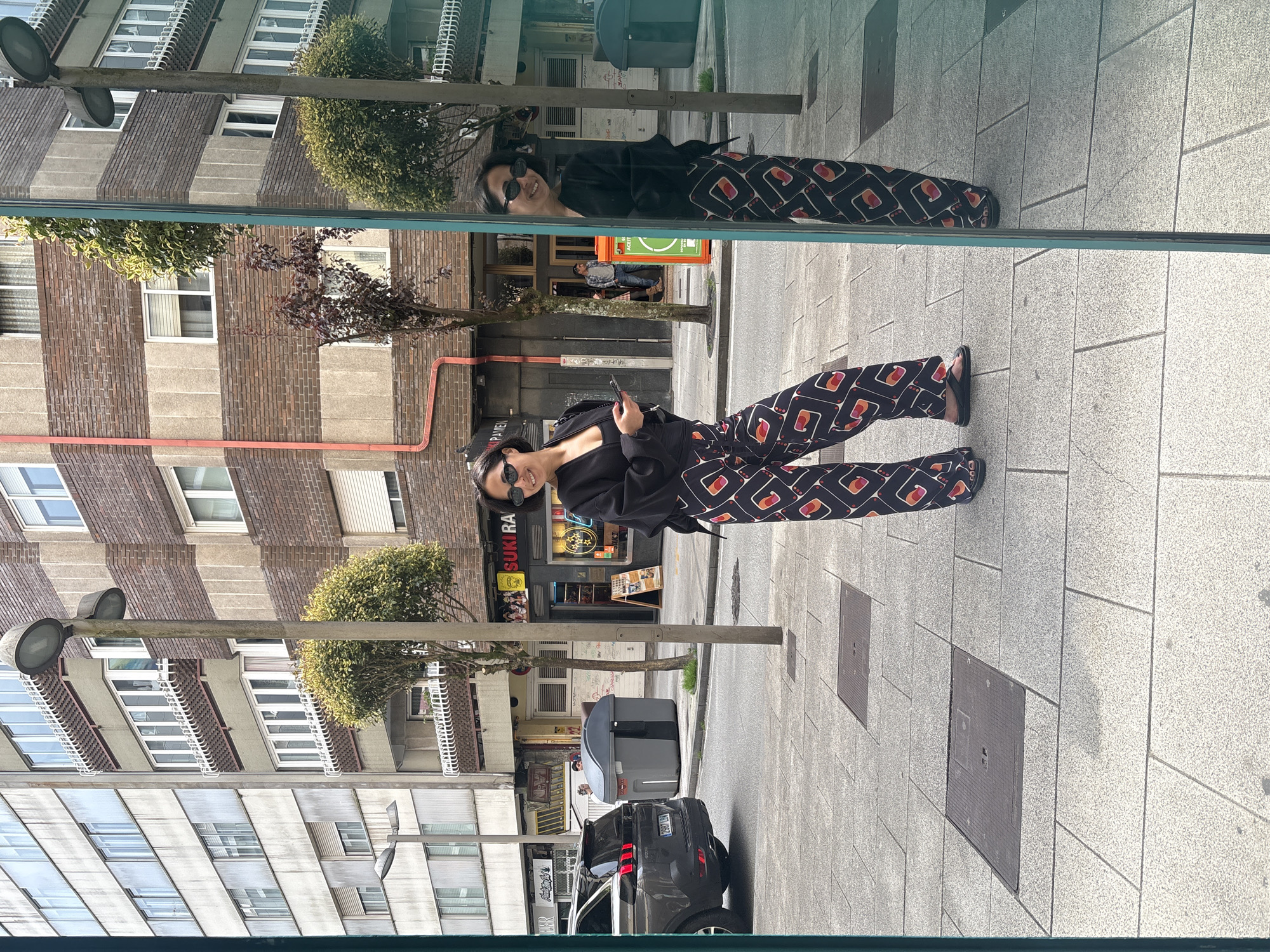 A woman in sunglasses, a black top, and patterned pants stands on a tiled sidewalk, holding a phone. She is captured as a reflection in a glass window, revealing a cityscape behind her. The reflection shows her mirrored image. The real-world backdrop features buildings, including a multi-story structure with striped brickwork, and street-level establishments like 'SUKIRA.' A dark-colored car with red taillights is parked nearby. The scene takes place in Santiago de Compostela, Spain, as inferred from local establishments and architectural style.