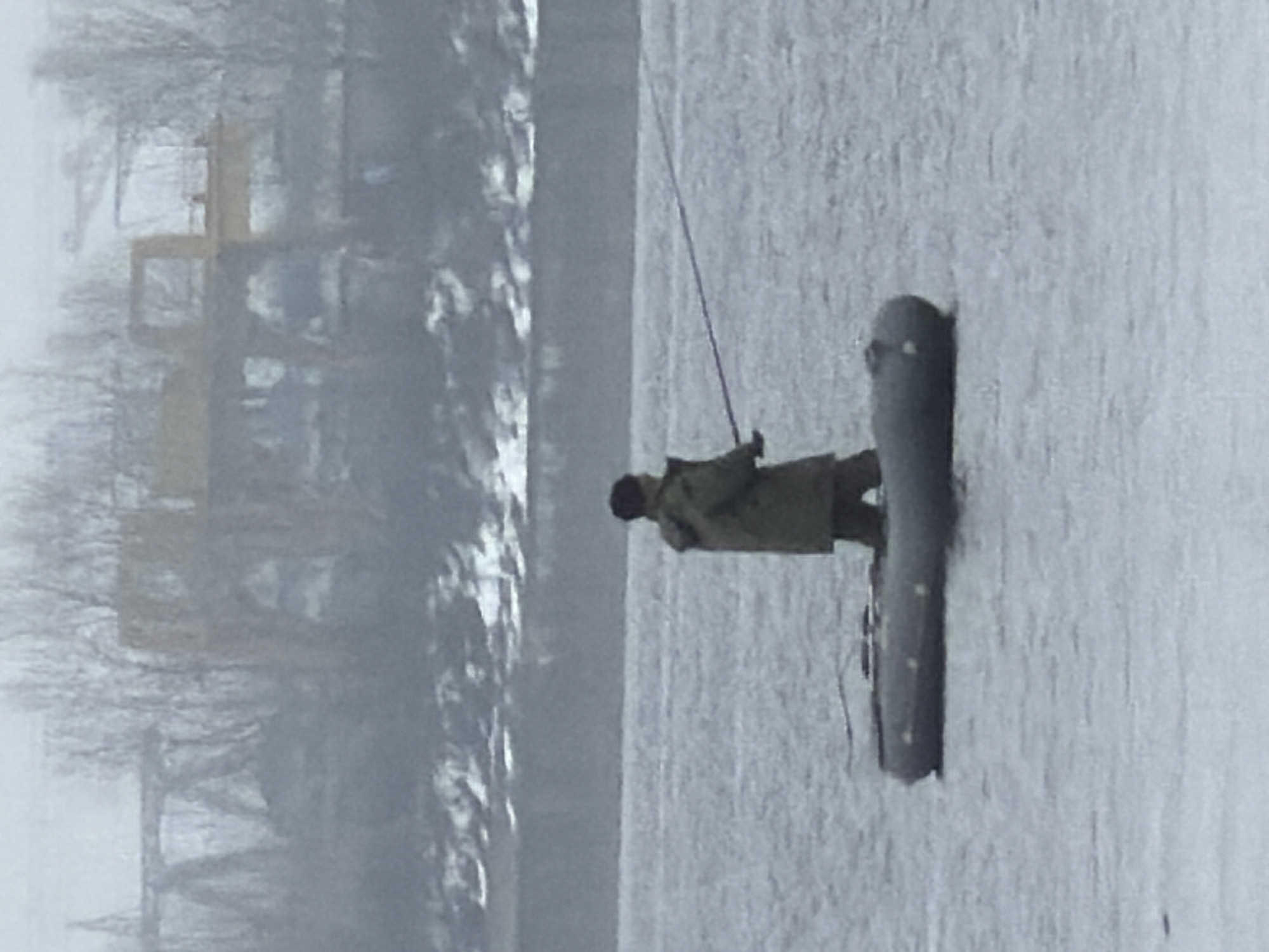 The image depicts a person ice fishing on a frozen body of water. The person is standing on a dark-colored sled-like object and holding a fishing rod. The person is wearing dark clothing, including a hat and a jacket. The background shows a blurry cityscape with bare trees and a yellow construction crane. Based on the visual details, this image appears to be taken in the city of Chaikovsky, Russia, during winter, and the person is ice fishing on the Kama River.