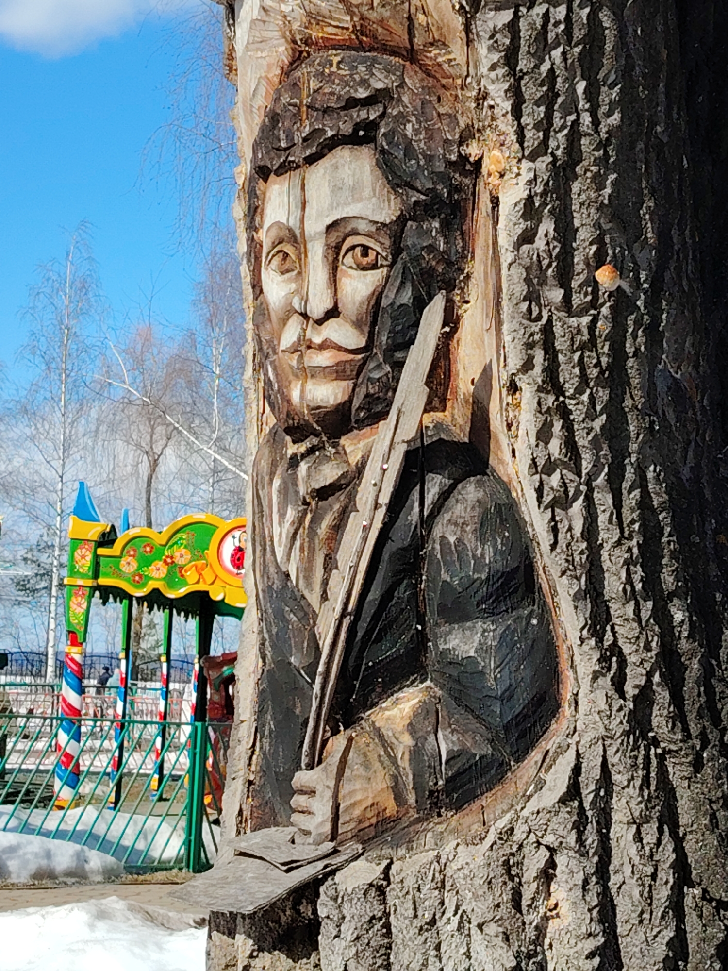 A tree carving of a man's bust is prominently featured on a tree trunk. The carving is detailed with the man wearing a dark suit and holding a writing utensil. The background shows a playground with colorful structures, suggesting a public park. Patches of snow are visible on the ground, indicating it's either winter or early spring. The location is likely Chaikovsky, Russia, as requested.