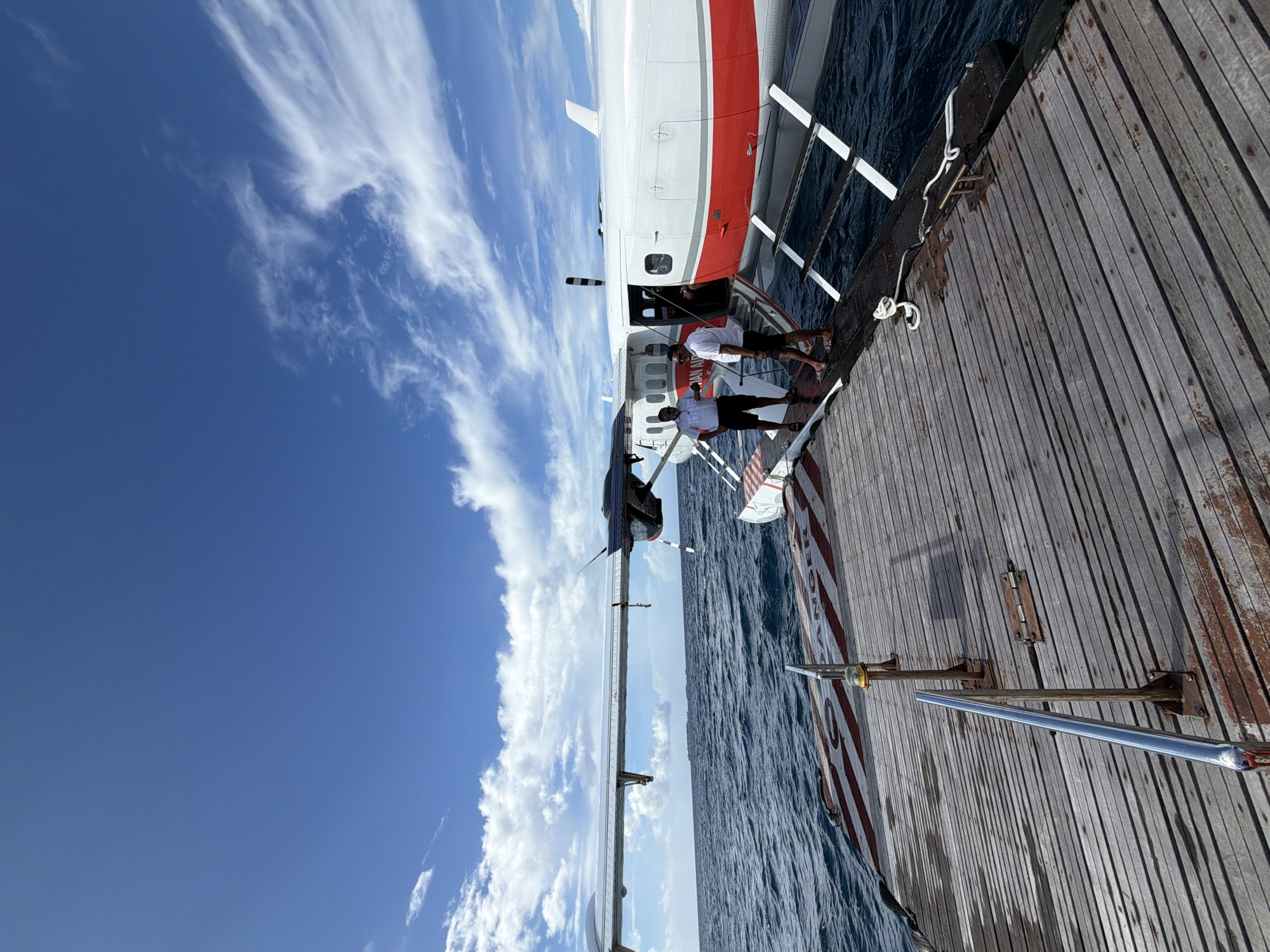 A seaplane, identified by the text "Malian Air" on its side, is docked at a wooden pier in Halaveli, Maldives. Two men, dressed in white shirts and dark shorts, stand on the pier near the seaplane's entrance, appearing to be assisting passengers or crew. The seaplane has a white fuselage with a red stripe along its lower section. It rests on floats in the ocean, which appears choppy with moderate waves. The sky is blue with scattered white clouds. The wooden pier is weathered and shows signs of wear. A red and white striped pattern is visible on the pier, potentially indicating a safety marking. The overall scene depicts a transportation hub in a tropical island setting.