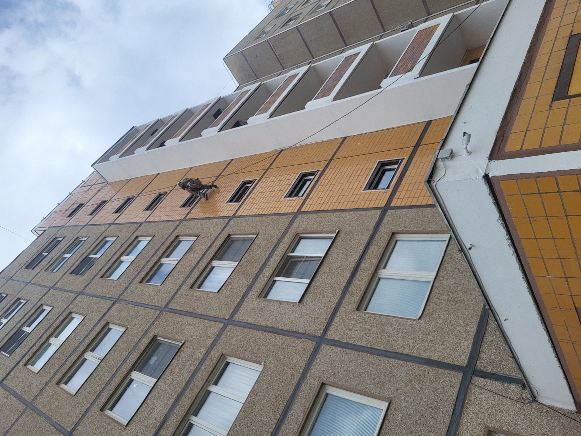 The image shows the exterior of a tall, multi-story building in what is identified as Chaikovsky, Russia. The building has a distinctive architectural style, with a combination of textures and colors on its facade. The lower levels are clad in a rough, textured material, divided into sections by dark gray or black trim. Windows are spaced regularly across this surface. Above this section, there is a band of yellow-orange tiling, with smaller, rectangular windows. Above this section, there are balconies. A man is suspended from the side of the building by ropes.