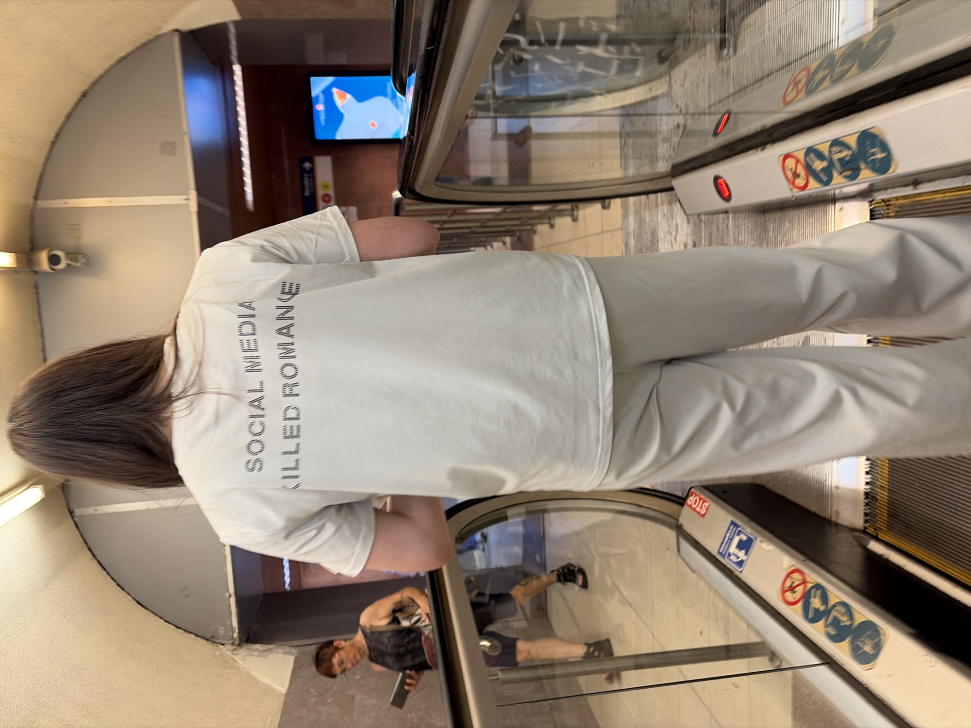 In an urban setting, possibly a subway or transportation hub, a person stands facing away from the camera on an escalator. The individual is wearing a light-colored t-shirt with black text on the back that reads "SOCIAL MEDIA KILLED ROMANCE" in all caps. They are also wearing matching light-colored pants. The escalator has metal railings and glass panels. To the left, another person with short hair and glasses is visible, looking down at a tablet. They are wearing a dark sleeveless top and shorts. The environment features a curved ceiling, overhead lighting, and advertisements on a screen in the background. Various safety stickers and signs are present on the escalator.