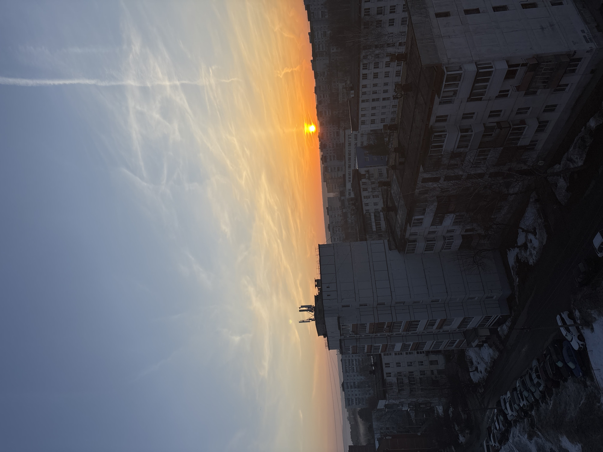 The image captures a sunset view over a densely populated urban area. The setting sun, a bright orange disc, dominates the horizon between rows of tall buildings. Above the sun, the sky is a blend of orange and yellow. Further from the sun, the sky transitions to a pale blue color. The sky is filled with thin, wispy clouds, and what appears to be a contrail cuts horizontally across the upper left portion of the image. The buildings are characterized by their uniform design with many windows and a gray color. Below the buildings, there is an area with cars parked along a street. The location is determined to be the city of Chaikovsky, Russia due to the location on the image file name.