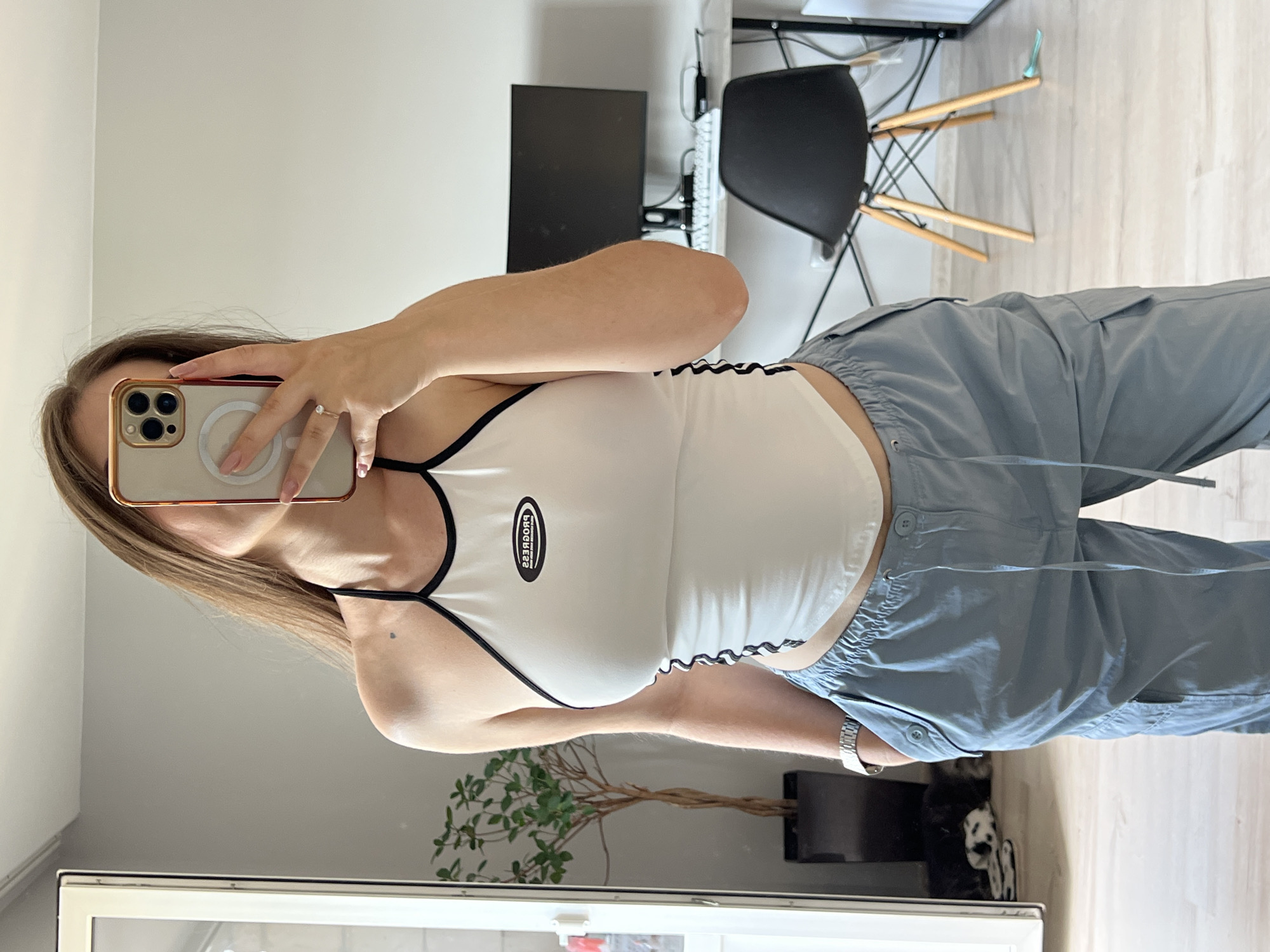 The image shows a woman taking a selfie in a room. She is wearing a white halter top with black trim and light blue cargo pants. She is standing in front of a mirror, with a phone in her hand. In the background, there is a desk with a computer monitor, a chair, and a plant. A dog is visible sleeping on the floor to the left of the frame. The overall setting appears to be a home office or living space. The location is not specified in the image, so it cannot be determined if it is in Ternopil, Ukraine.