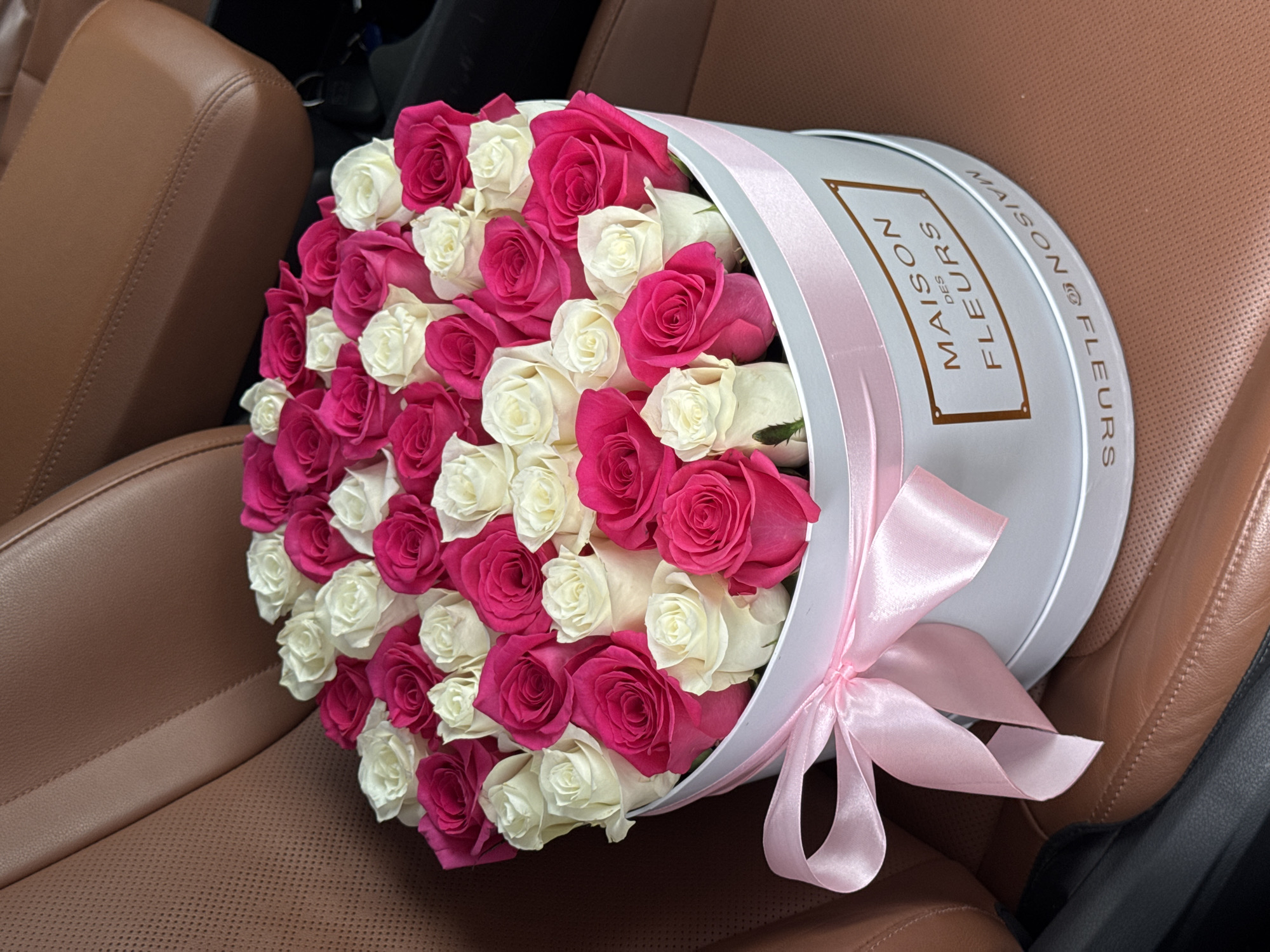 A close-up, eye-level shot shows a white round floral box filled with numerous pink and white roses. The box is tied with a pink satin ribbon. A gold-colored plaque on the side of the box reads "MAISON DES FLEURS." The box is situated on a brown leather car seat. The background is dark and out of focus, suggesting the interior of a vehicle. The image is taken indoors.