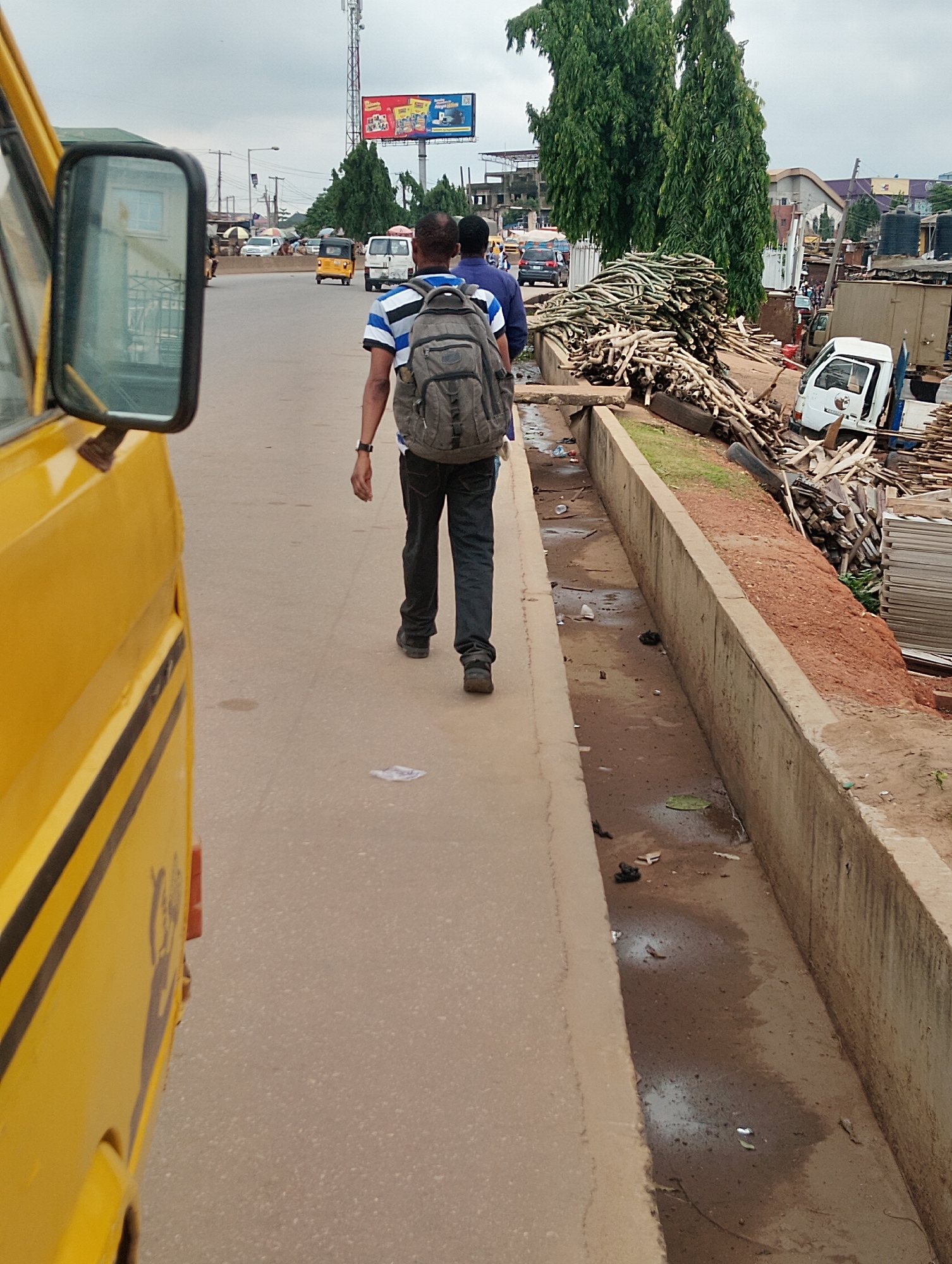 A person walks on a paved sidewalk next to a yellow bus. The person, wearing a blue and white striped shirt and dark pants, carries a grey backpack. They are walking away from the camera, on a street in Agege, Nigeria. The street is lined with trees and buildings. A large pile of cut bamboo or sugarcane stalks is visible on the right side of the image, next to a concrete drainage ditch filled with murky water and debris. Several vehicles, including a white van and a yellow auto-rickshaw, are in the background. The sky is overcast.
