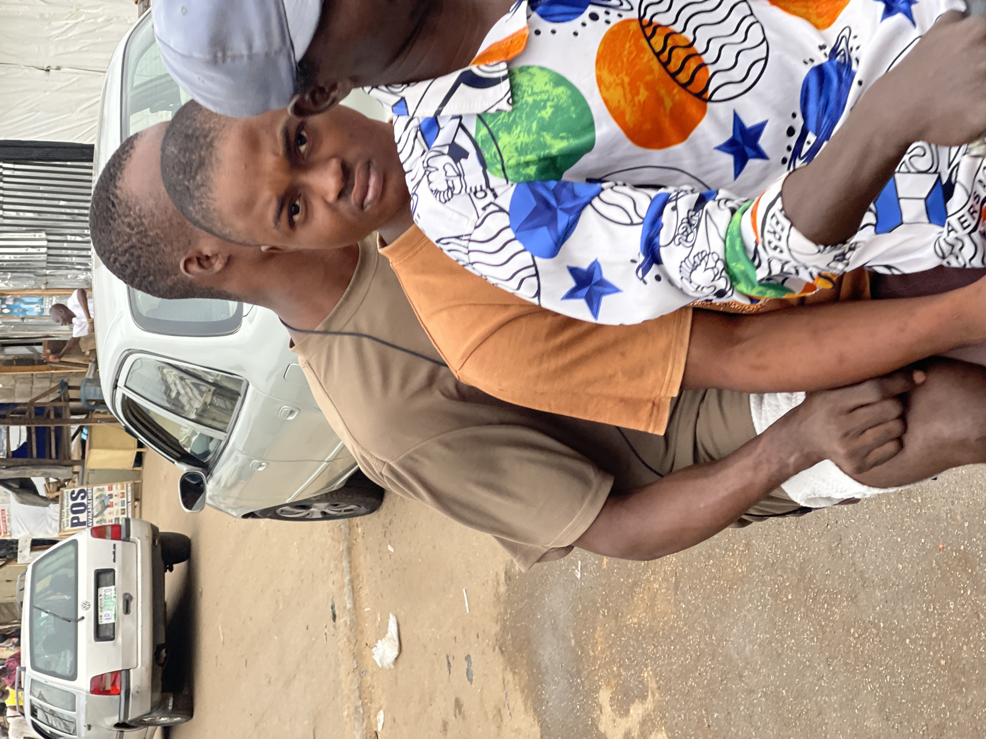 The image captures a scene in Kuchingoro, Nigeria, focusing on two males in close proximity. One male, wearing a brown t-shirt, is partially visible behind the other. The second male wears a white shirt patterned with blue stars, orange circles, and green shapes. The background includes a silver car and a white car with a visible license plate that reads "Lagos". A sign with the word "POS" is also visible, likely indicating a point-of-sale terminal nearby. The ground is a concrete surface scattered with small pieces of debris. The overall scene suggests an urban environment.