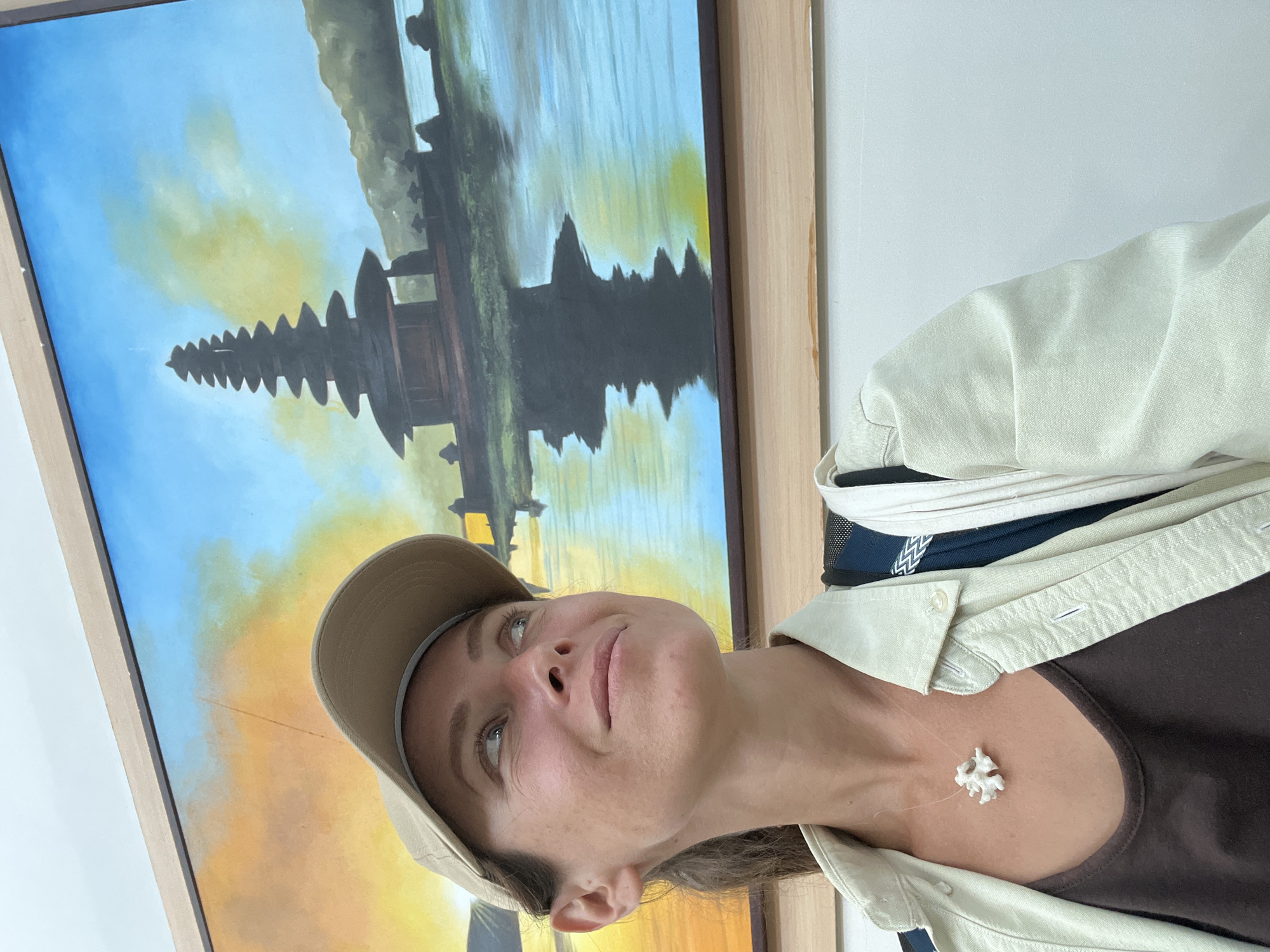 A woman wearing a beige baseball cap, a beige collared shirt over a dark brown tank top, and a white coral necklace poses for a selfie in front of a painting. The painting depicts a multi-tiered pagoda structure on a body of water, with a colorful sky. The woman looks up and to the right with a slight smile. She has a backpack strap over her right shoulder. The background wall appears to be white.