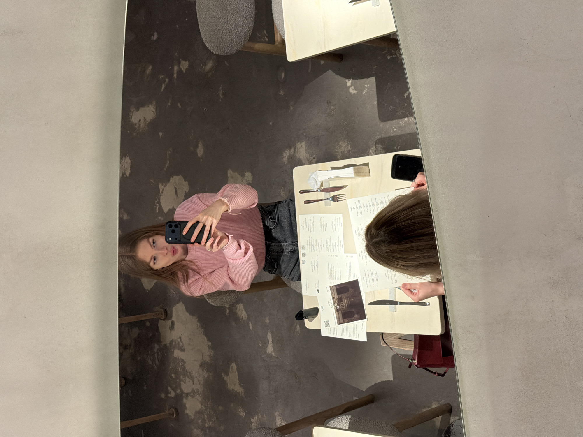 The image shows a woman taking a selfie in a mirror. The reflection shows a restaurant scene: two women sitting at a table, menus, cutlery, and a purse. The restaurant floor is patterned grey. While the location of the restaurant cannot be determined from the visual content alone, this additional information cannot be confirmed without more specific visual clues or contextual data.
