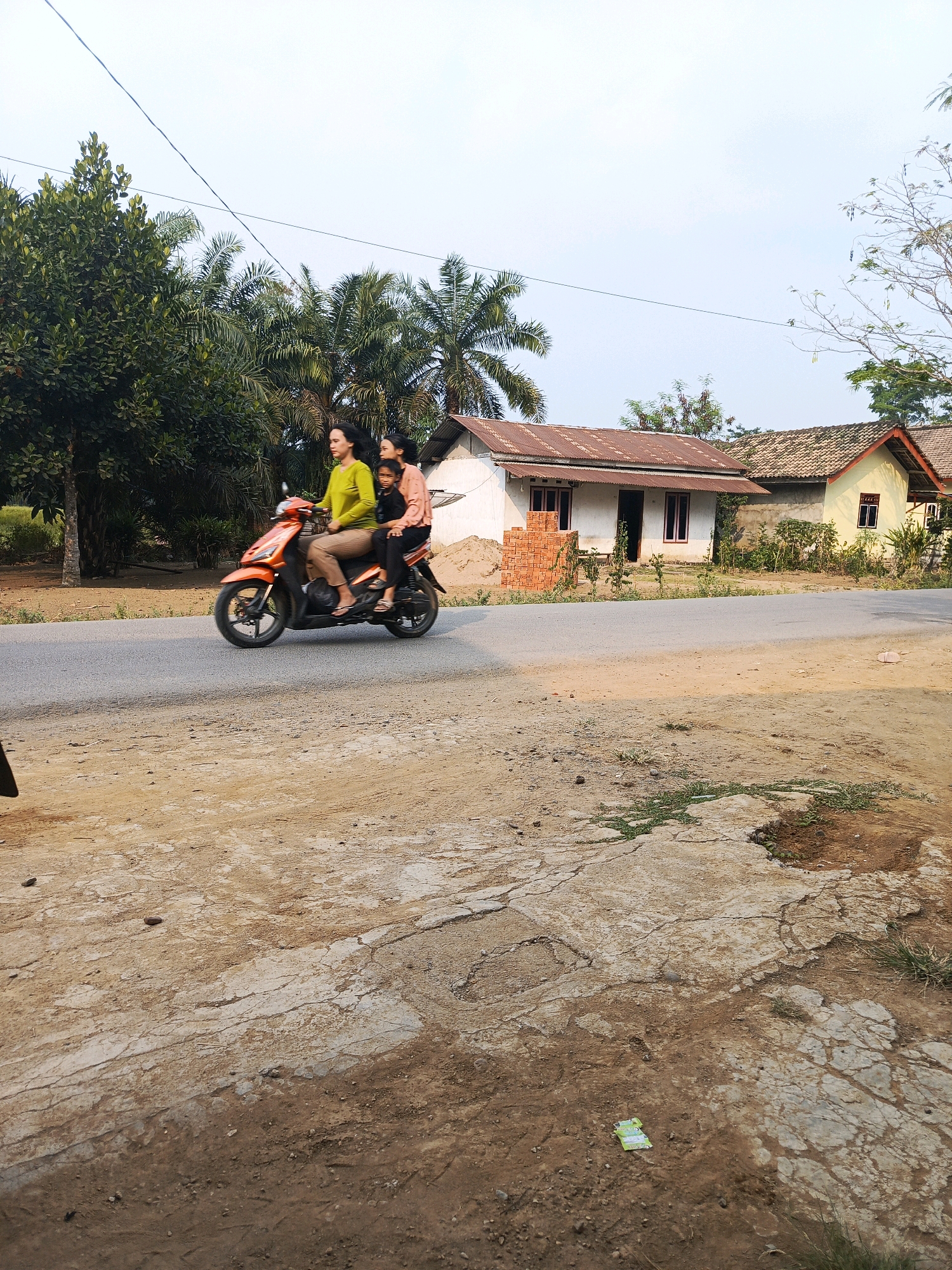 In Megang Sakti I, Indonesia, an orange scooter navigates a paved road, carrying three individuals. A woman in a bright green shirt and tan pants drives, a child in a peach-colored shirt sits behind her, and another child in a dark shirt is positioned behind the driver. The scooter is captured in motion, leaning slightly as it turns. To the right of the road, a row of houses with tin and tiled roofs is visible, surrounded by lush green trees and palm trees. The foreground of the image shows a dirt and cracked concrete ground, with sparse patches of grass. The sky is clear and bright, suggesting daytime.