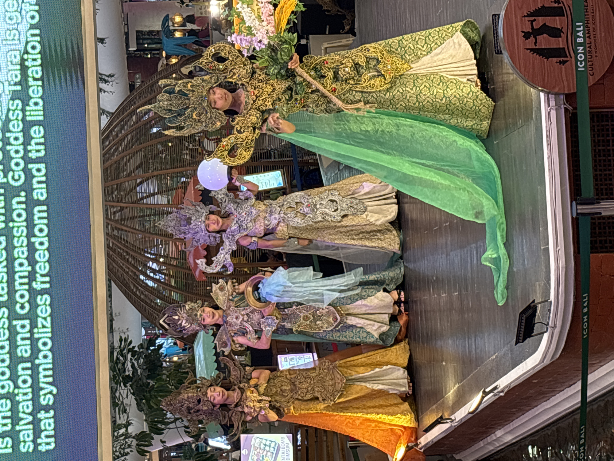 Four young women in elaborate traditional costumes are standing on a stage. The women are wearing ornate headdresses and ornate, long gowns. The woman on the far left wears a gold and orange gown. The second woman from the left wears a green and gold gown and holds a circular object. The third woman from the left wears a purple and silver gown. The woman on the far right wears a green and gold gown, with a long green sash draped around her. She is holding a bundle of green foliage. The stage is made of dark wood and is raised above a body of water. There is a large screen behind the women displaying text. In the foreground, there is a sign with the words "ICON BALI CULTURAL AMPHITHEATER" printed on it. The overall impression is that of a cultural performance. The image was taken at ICON Bali, a cultural amphitheater located in the city of Denpasar, Indonesia.