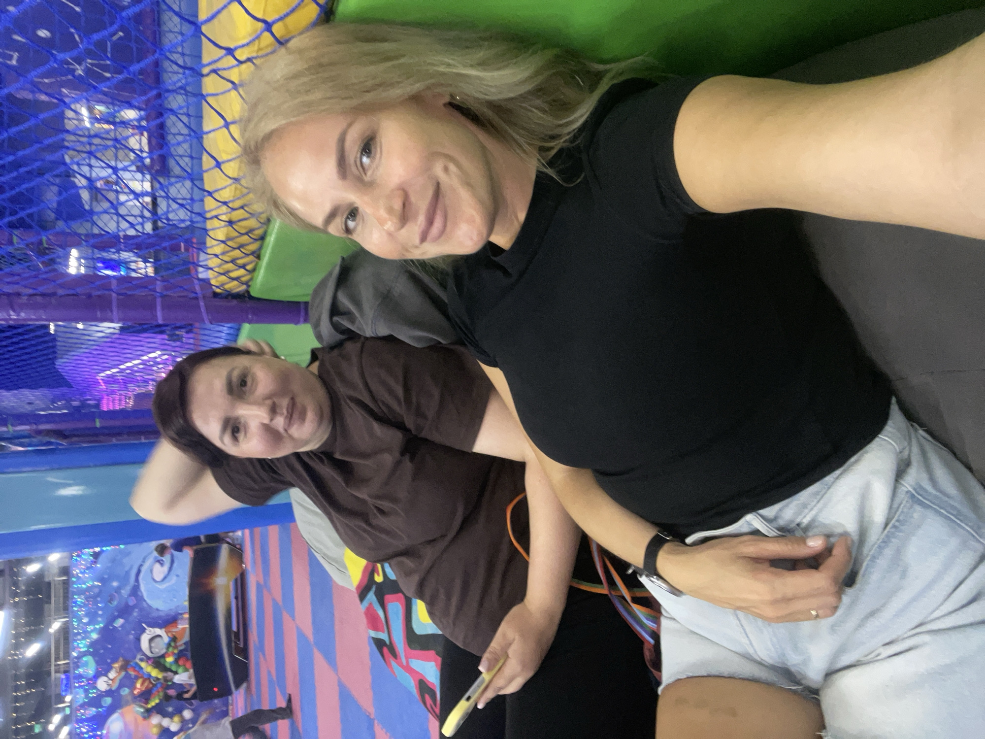 Two women are posing for a selfie. The woman in the foreground has blonde hair, is wearing a black t-shirt and light blue denim shorts. The woman in the background has dark hair, is wearing a brown t-shirt, black pants, and is holding a cell phone. They are indoors, possibly in a play area or family entertainment center. The background includes colorful mats, a netting structure, and a mural depicting space-themed elements. The location is the city of Sochi, Russia.