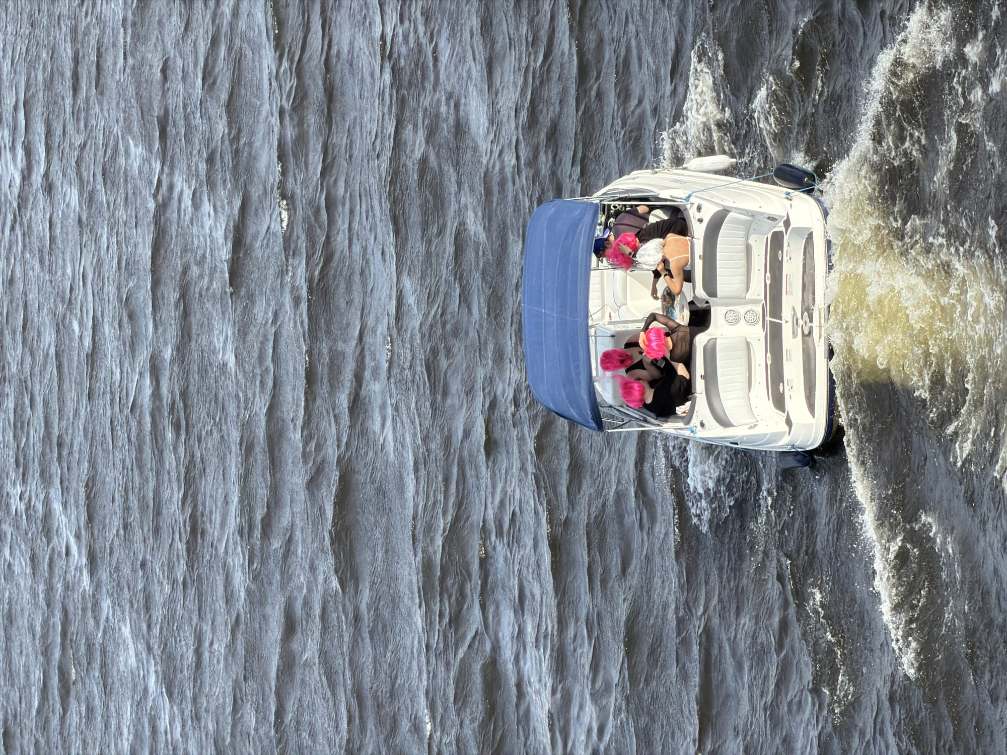 A white motorboat with a blue Bimini top is depicted in motion on dark, rippled water, situated in the city of Saint Petersburg, Russia. The boat is viewed from a high-angle perspective, showing its stern and a distinct foamy wake trailing behind. There are at least six individuals on board, seated within the cockpit area. The majority of the occupants are wearing bright neon pink wigs, while one person towards the rear right side of the boat is wearing a white wig. They appear to be engaged in social interaction, with some gathered around a small table that holds unidentified items. Two black fenders are visible attached to the starboard side of the boat. The water surface exhibits moderate choppiness, reflecting ambient daylight.