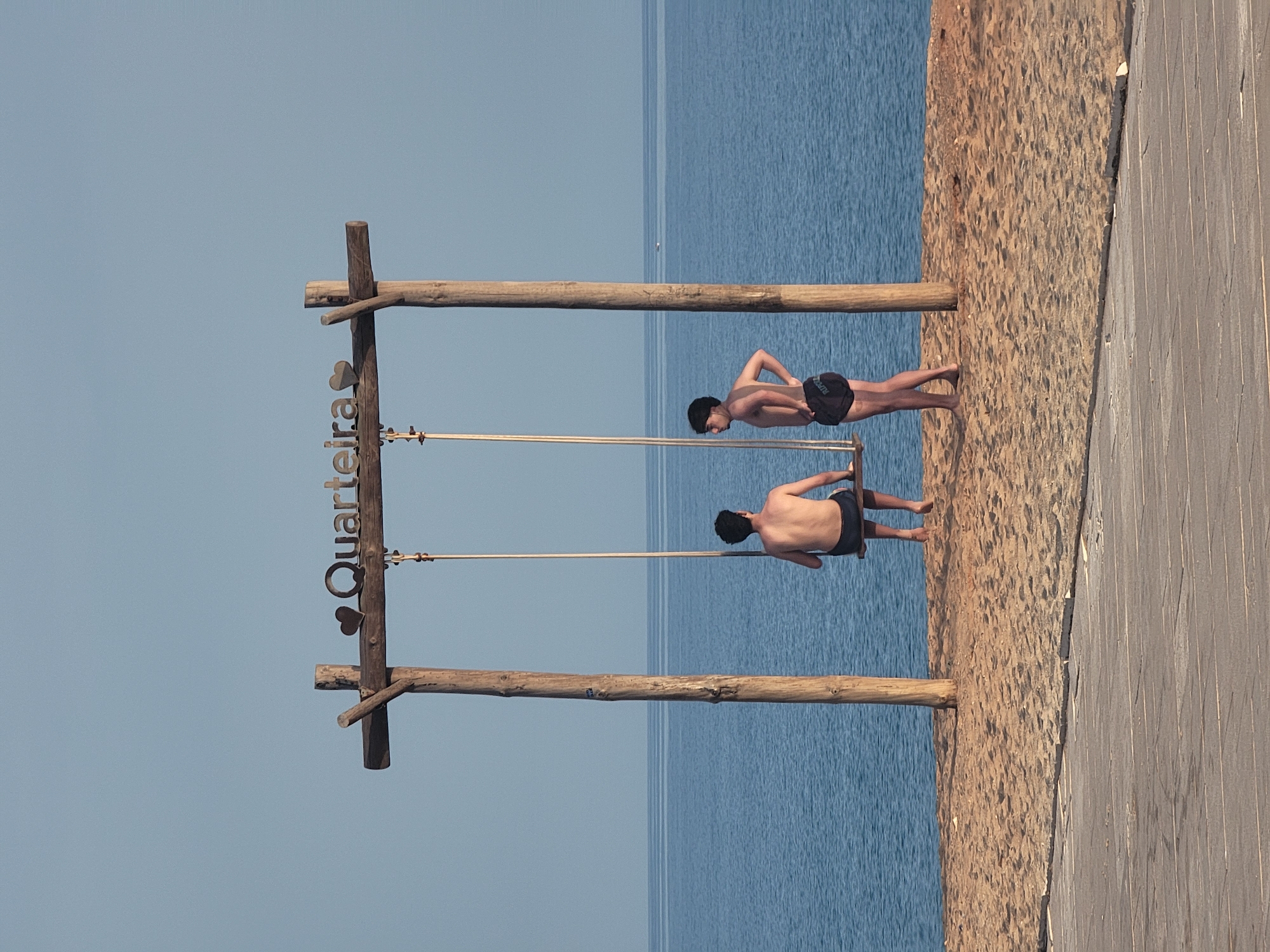 The image shows two young men on a beach in Quarteira, Portugal, with a wooden swing set in the foreground. The swing set has the word "Quarteira" with two hearts above it, crafted from metal. The sea is visible in the background, calm and blue under a clear sky.
One man is sitting on the swing, facing away from the camera, wearing dark swim shorts. The second man is standing to the right of the swing, also facing away, wearing black swim shorts. His left hand is on his hip, and his right hand is slightly bent. Both men are bare-chested.
The beach is made of light brown sand, and in the very foreground, a wooden boardwalk or walkway is visible. The overall scene suggests a relaxed, daytime atmosphere by the sea.
