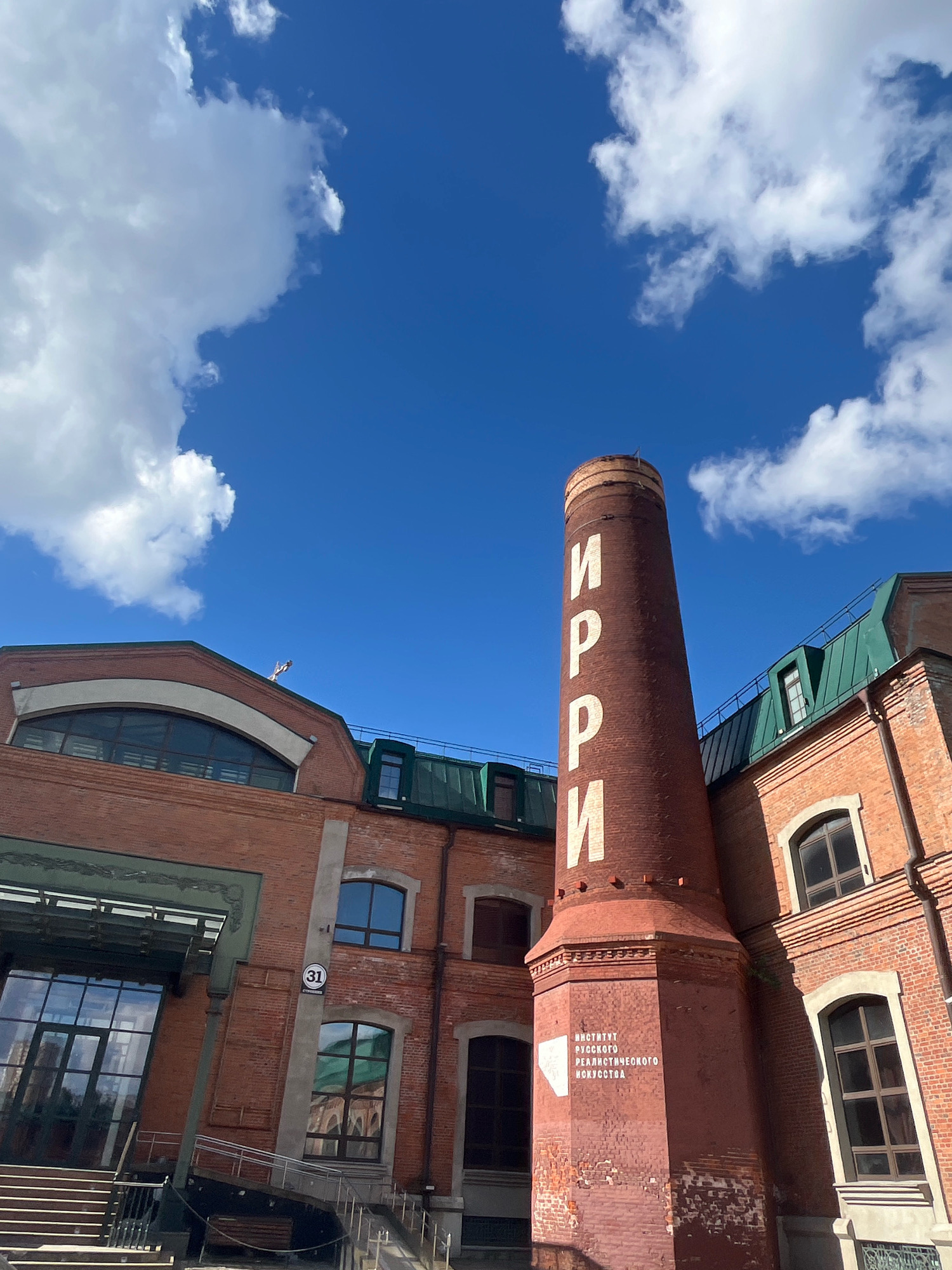 The image captures a view of the "Institute of Russian Realist Art" in Moscow, Russia, on a bright, sunny day with a vivid blue sky and scattered white clouds. The primary focus is on a tall, red-brick chimney with white Cyrillic lettering. The letters "И", "Р", "Р", "И" are prominently displayed vertically on the upper portion of the chimney. Lower down on the chimney, smaller white text reads "ИНСТИТУТ РУССКОГО РЕАЛИСТИЧЕСКОГО ИСКУССТВА" (Institute of Russian Realist Art).
To the left of the chimney, a large brick building with a green-tiled roof is visible. This building features large arched windows and a modern glass entrance with a covered walkway. A circular sign with the number "31" is affixed to the brick facade. Other brick buildings with green roofs and arched windows are also visible. The architecture suggests a repurposed industrial or historical complex. The perspective is from a low angle, looking upwards towards the sky. No people are visible in the scene. The overall impression is one of historical architecture set against a clear, expansive sky.