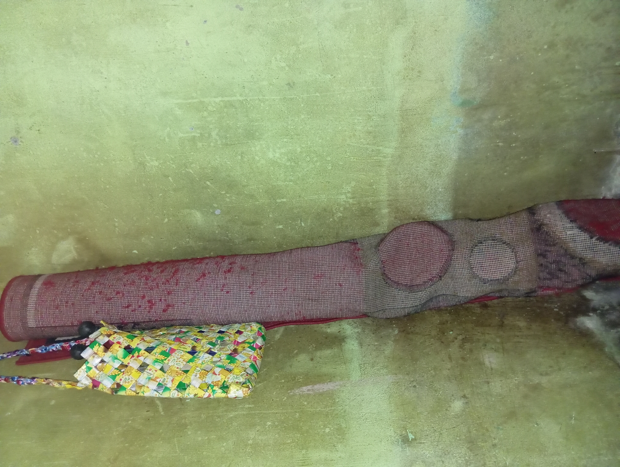 A woven bag made of colorful plastic is hanging on a light green, textured wall in Rejosari, Indonesia. Next to the bag, a rolled-up mat with a red and dark pattern is leaning against the wall. The mat appears to be made of a woven material with a coarse texture. Below the mat, a section of the same material is visible, displaying a circular pattern within a grid. The wall shows signs of wear and tear, with subtle discoloration and a rough surface. The lighting is subdued, casting soft shadows.