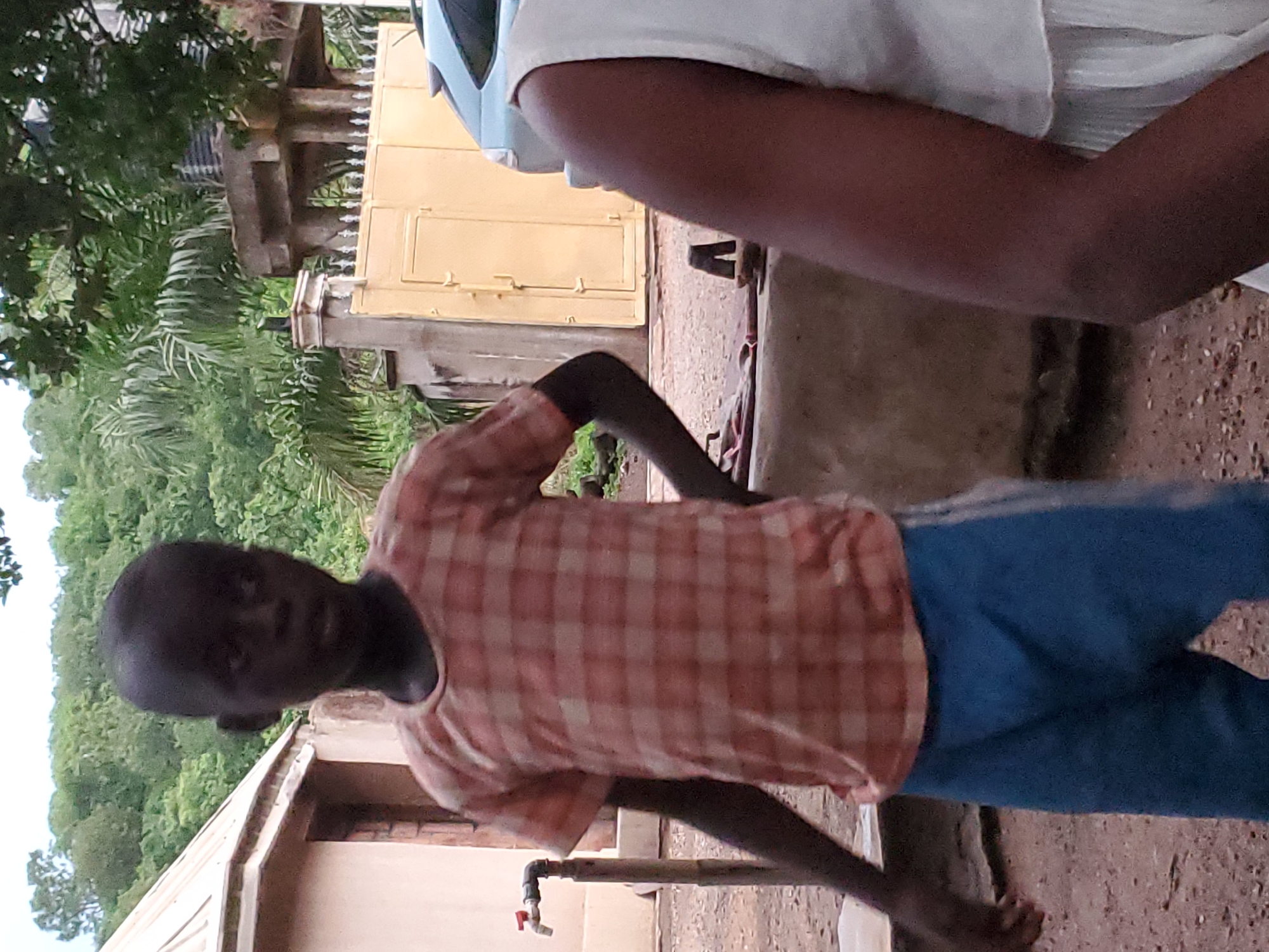 A boy stands near a water tap outside a building in Mararaba, Nigeria. He wears a plaid shirt and blue pants. Another person, partially visible, is nearby. Vegetation is in the background. A building with a yellow door is also visible. The ground is dirt.