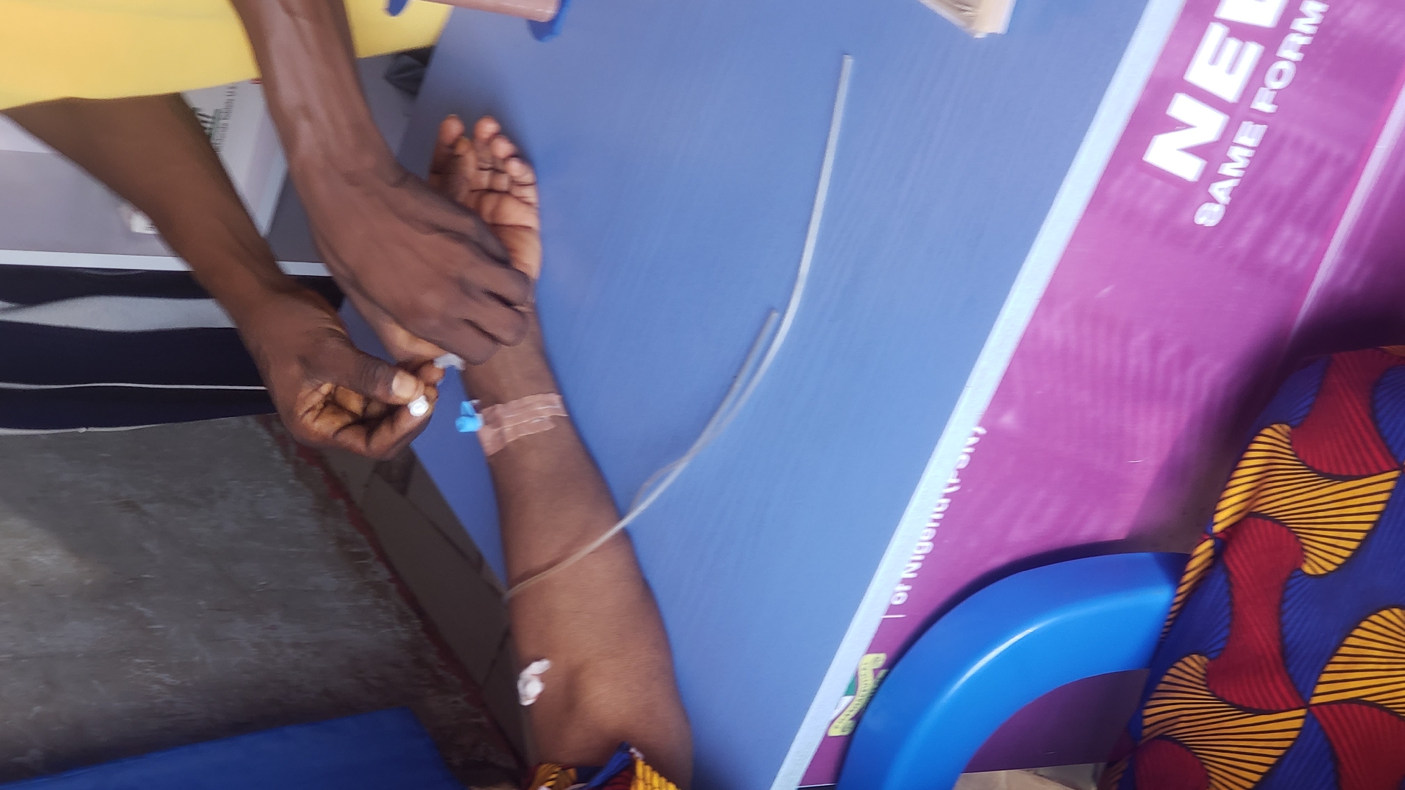 The image depicts a close-up view of a medical procedure involving a person's arm. A medical professional, whose hands are visible, is attending to an intravenous (IV) line inserted into the forearm of another person. The IV line is secured with medical tape. A blue cap is attached to the IV connection. The scene appears to be set indoors, with a blue surface and a portion of a purple sign with white text visible in the background. The text on the sign includes "NE," "SAME FORM," and "of Nigeria (P-ON)." The person receiving the IV is wearing a colorful patterned garment, predominantly blue, red, and yellow. The overall impression is one of a medical intervention taking place in a clinic or hospital setting. The lighting suggests daytime.