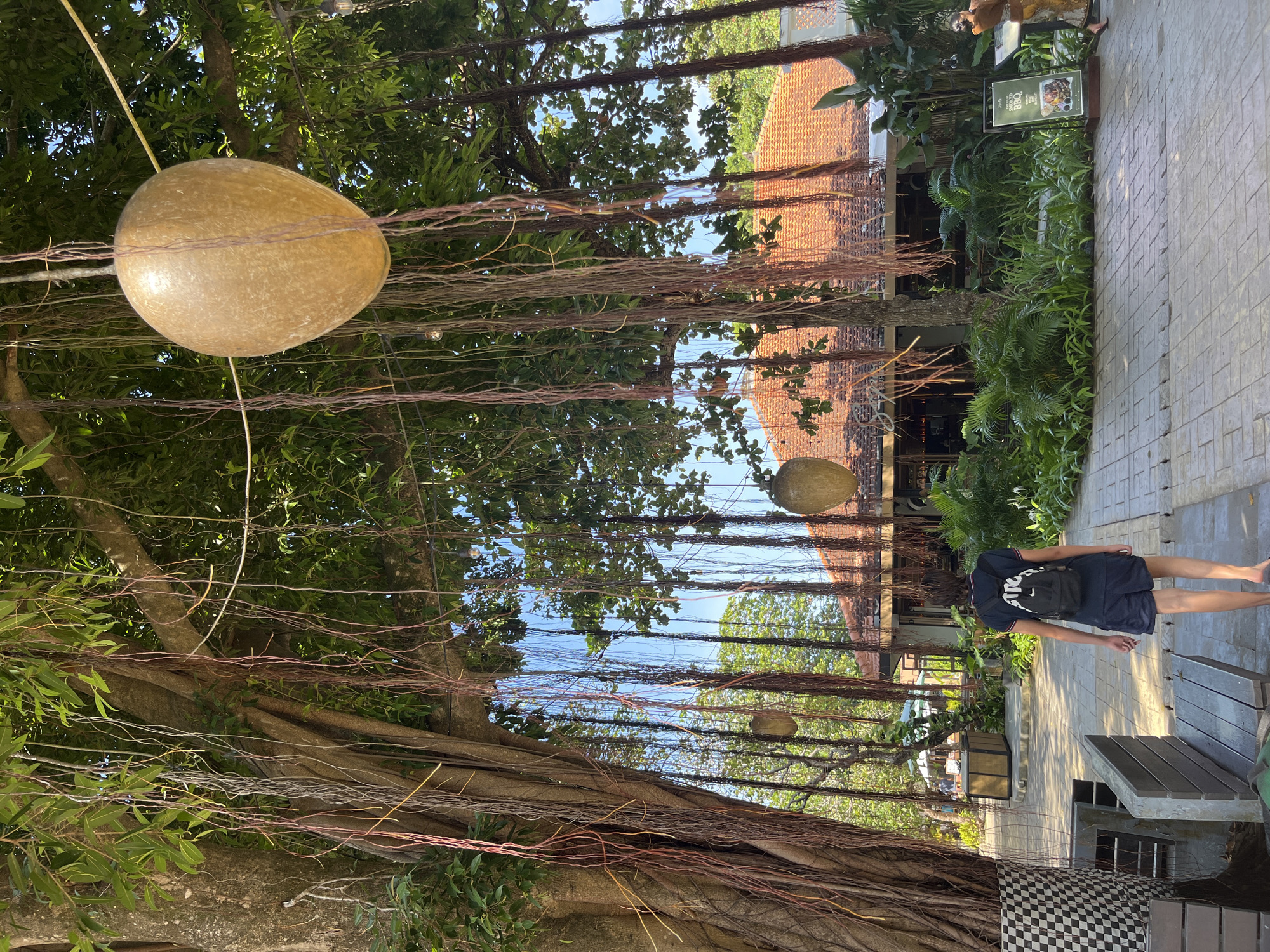 In Kuta Selatan, Indonesia, a young person with a black backpack walks away from the camera on a paved walkway. They are wearing a dark t-shirt and shorts. The scene is outdoors, under the canopy of a large tree with numerous hanging aerial roots. In the background, a building with a red tiled roof is visible, surrounded by lush green foliage. Several large, golden, teardrop-shaped lanterns are suspended from the tree branches. To the left of the walkway, there are wooden benches and a checkered patterned structure. A sign for "SMOKED BBQ" is placed near the foliage on the right side of the image. The sky is clear and blue.