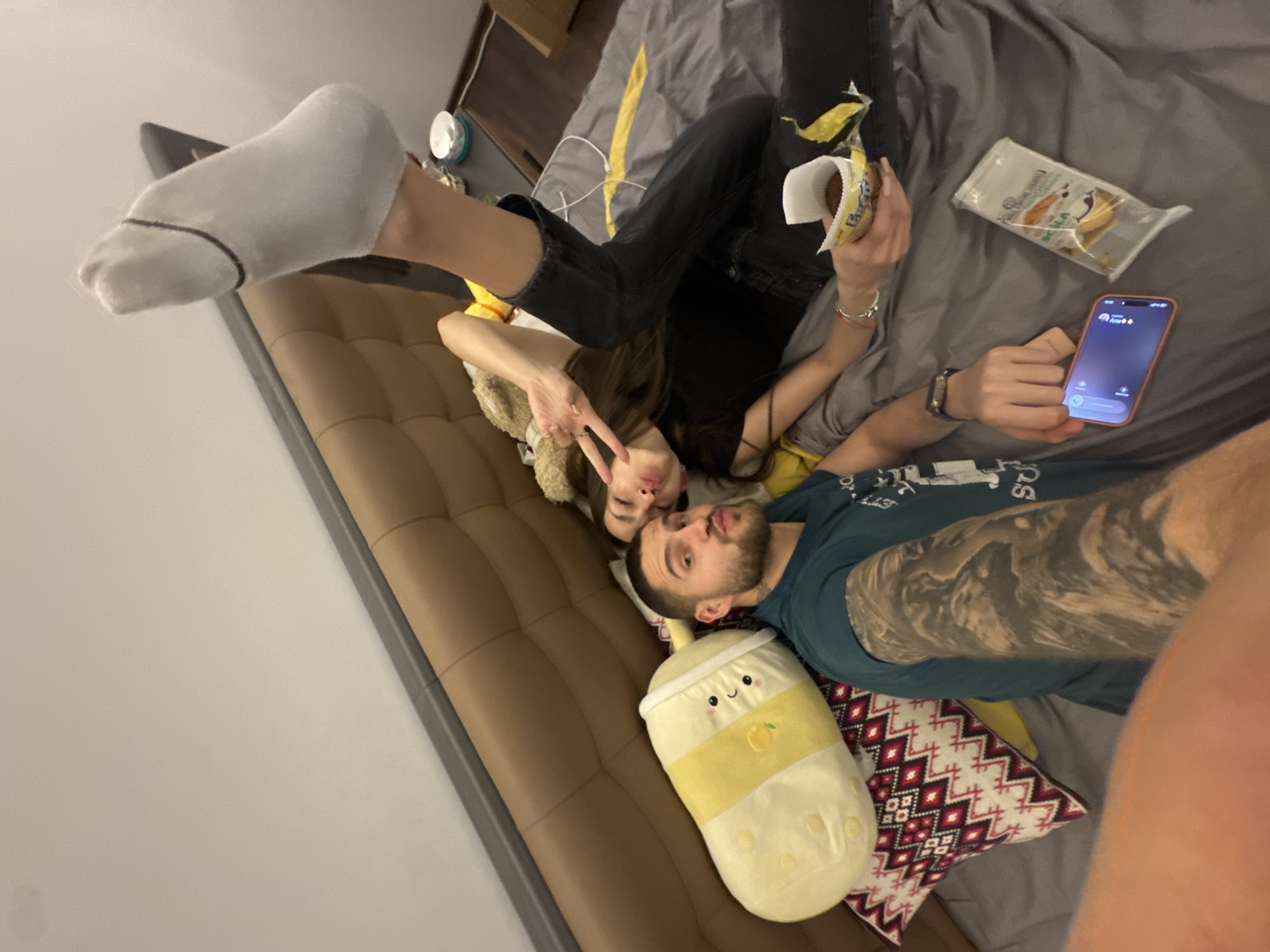 A couple is lying on a bed with grey sheets. The woman, wearing black jeans and a black top, is holding a snack in a white wrapper. She is making a peace sign with her hand. Her feet, with white socks, are raised. The man is next to her, wearing a blue shirt and a wristwatch. He is holding a mobile phone with an active phone call. A yellow boba tea pillow and a patterned pillow are nearby. There are snacks on the bed. The location appears to be indoors, likely in a home, in Sofia, Bulgaria, given the available visual information.