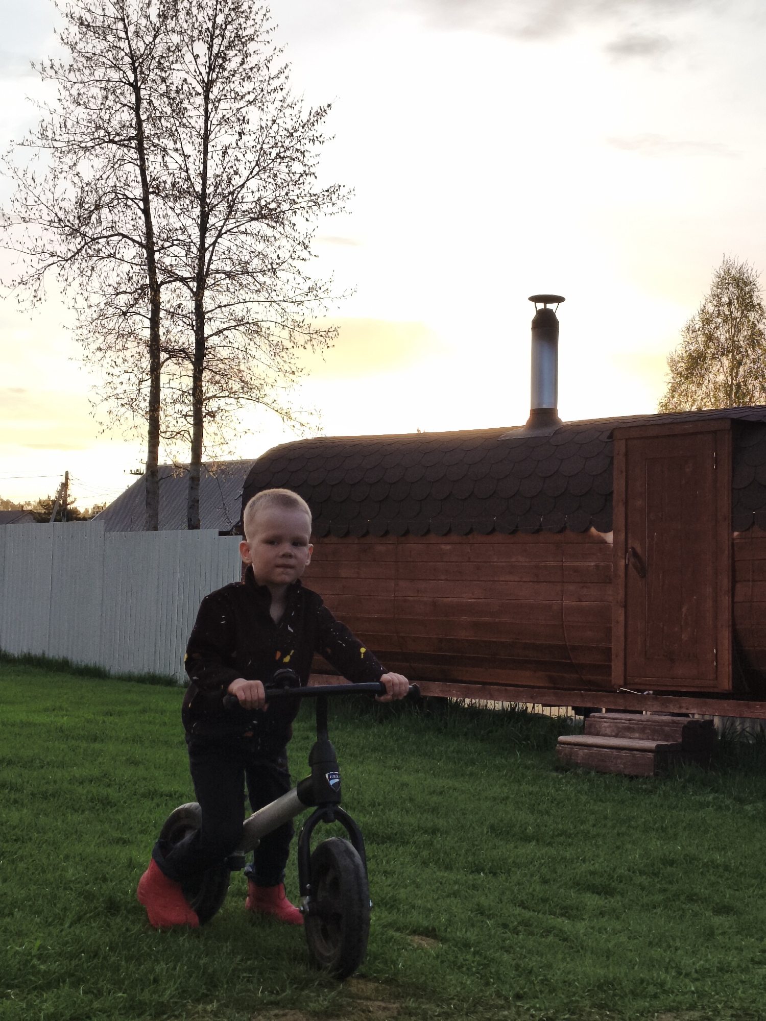 A young child is on a balance bike in a grassy area. The child is wearing a dark jacket and pants, with bright red boots. Behind the child is a small, dark wood structure, likely a sauna or shed, with a chimney extending from the roof. The area is enclosed by a white fence. There are bare trees visible in the background against a dusk sky. The location is likely Русский Вожой, Russia based on the request.