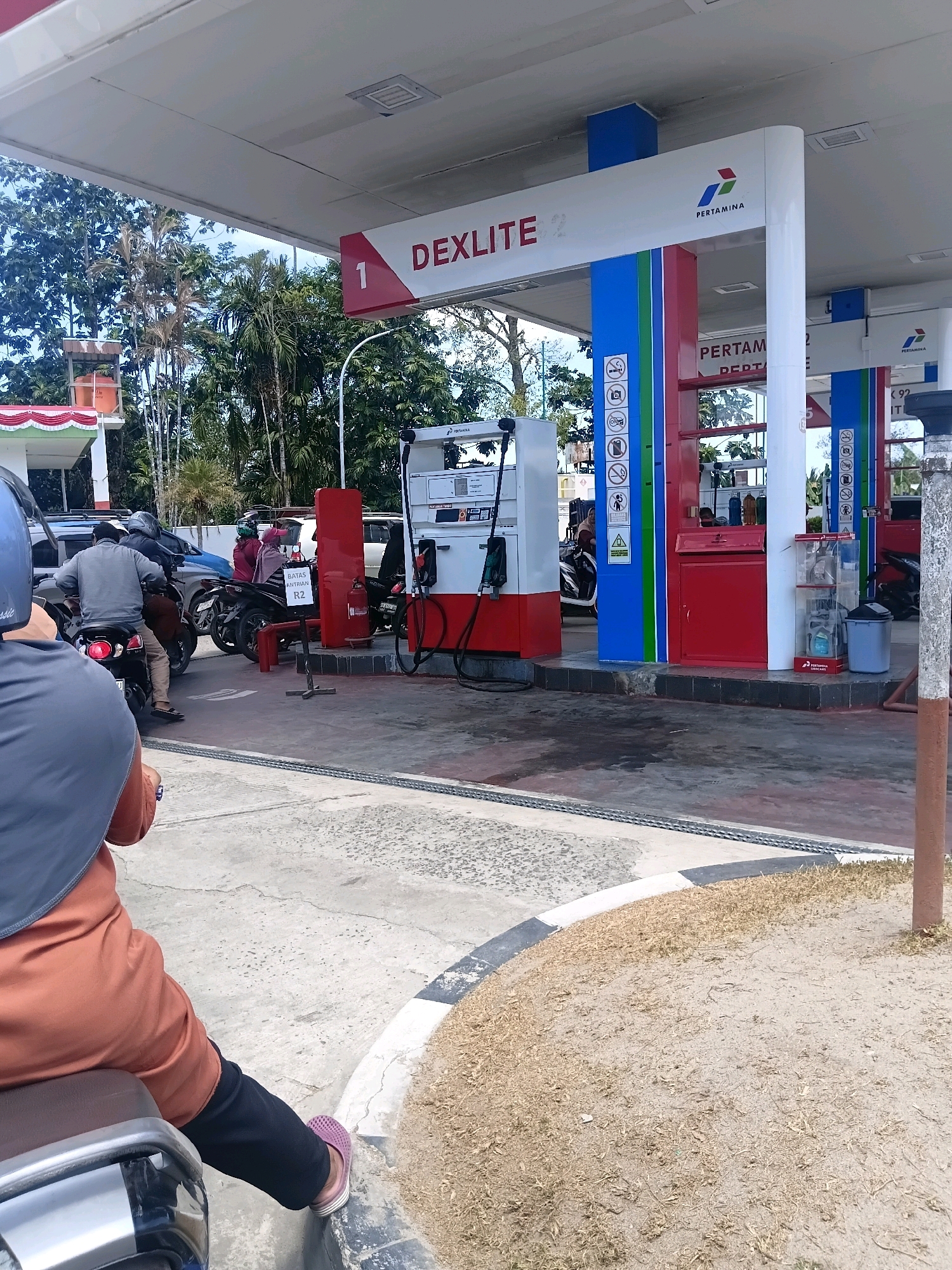 The image depicts a Pertamina gas station in Sungai Kapitan, Indonesia. In the foreground, a person is seated on a motorcycle, facing away from the camera, wearing a brown hijab and orange top. Another person on a motorcycle is visible further back, wearing a grey jacket and helmet. Several other motorcycles and vehicles are present at the gas station, with people in the process of refueling. The gas station features multiple pumps with the Pertamina logo and "DEXLITE" signage. The environment appears to be outdoors, with trees and vegetation visible in the background. The ground surface is paved concrete, with some areas of dry grass and sand. A red fire extinguisher is mounted on a pole. The scene suggests a typical day at a busy gas station.