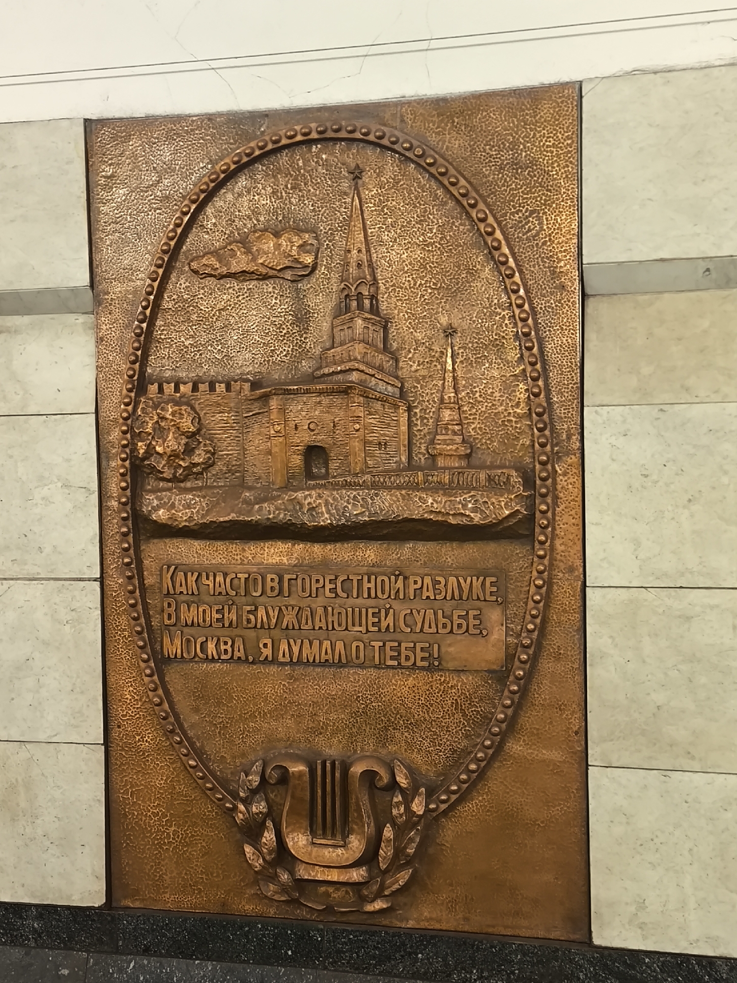 The image shows a copper-colored relief plaque mounted on a wall, likely in the Moscow metro. The plaque features an oval design containing a depiction of the Moscow Kremlin, a cloud, and a lyre surrounded by laurel branches. Inscribed in Russian within the oval is the text, which can be translated as "How often in sorrowful separation, in my wandering fate, Moscow, I thought of you!". The plaque is situated between sections of wall covered in white marble tiles.