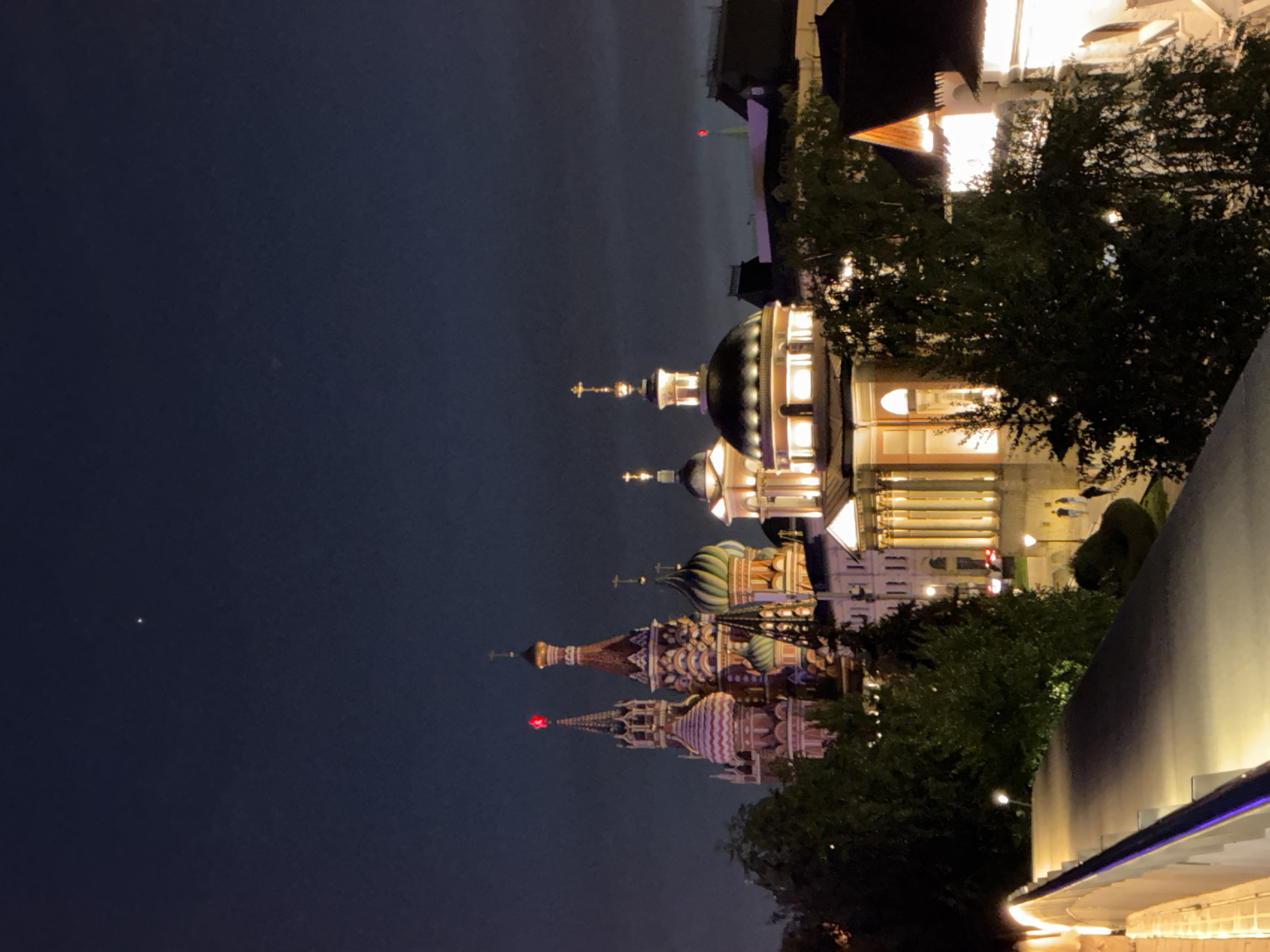 The image shows a night view in Moscow, Russia. Prominently visible are the illuminated domes of the Saint Basil's Cathedral and the Cathedral of Christ the Savior, indicating the location near the Red Square. The sky is dark, with one distinct light spot, likely a star or distant light. Trees are present, partially obscuring the view of the buildings. A structure with bright, linear lighting is visible at the bottom right.