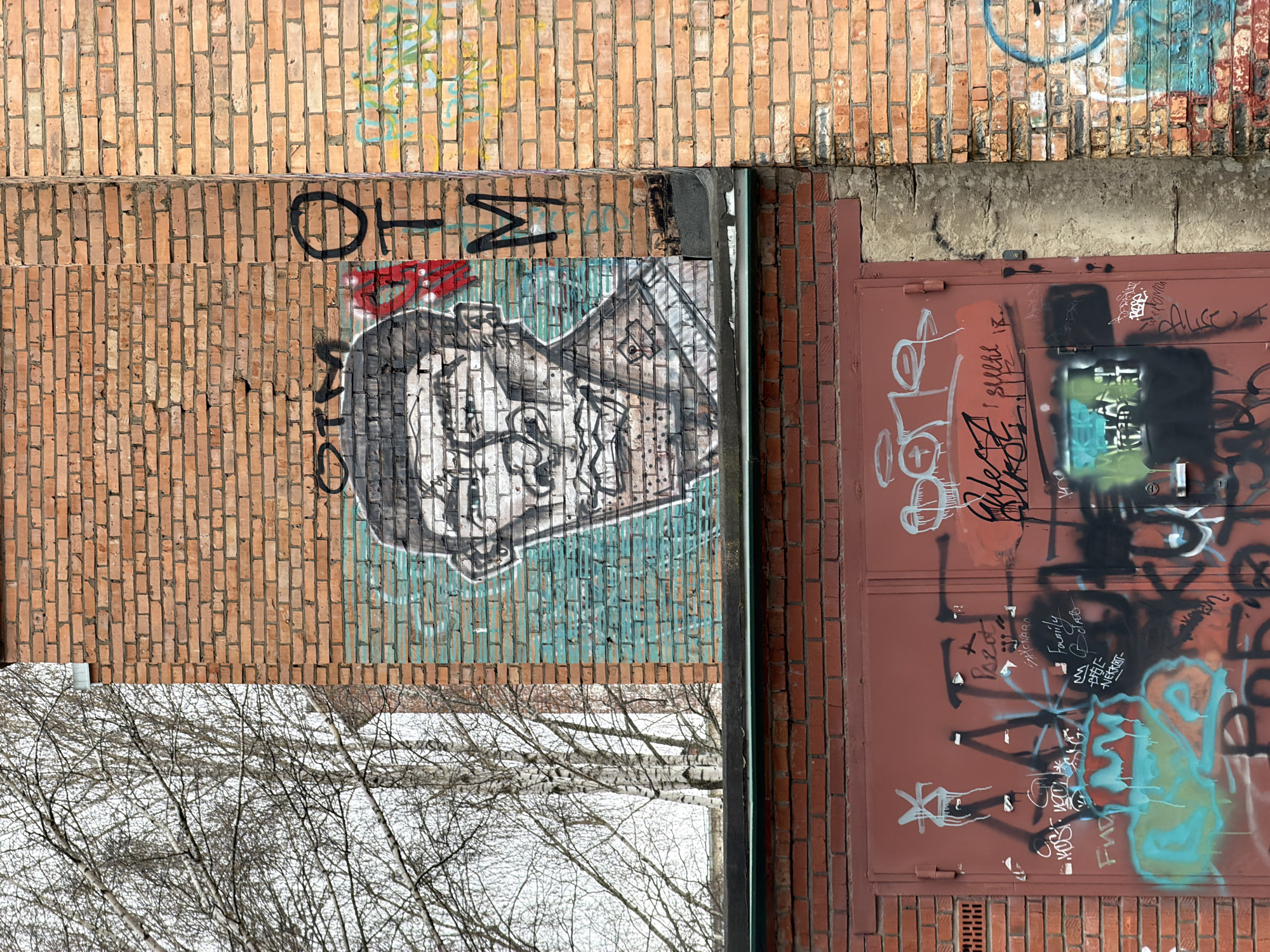 The image shows a section of a brick building in Votkinsk, Russia. The building has multiple layers of brickwork, with variations in color and texture. The upper sections are predominantly orange-red, while the lower sections exhibit a more aged, weathered appearance. Graffiti is visible on the brick surfaces, including what appears to be a portrait sketch in black and white with green accents. Other graffiti includes lettering and abstract designs. A section of the building has a metal door or panel, painted a reddish-brown, and heavily tagged with graffiti. In the lower portion of the image, bare tree branches are visible, suggesting it is winter or late fall. The background is a mixture of snow and grey.