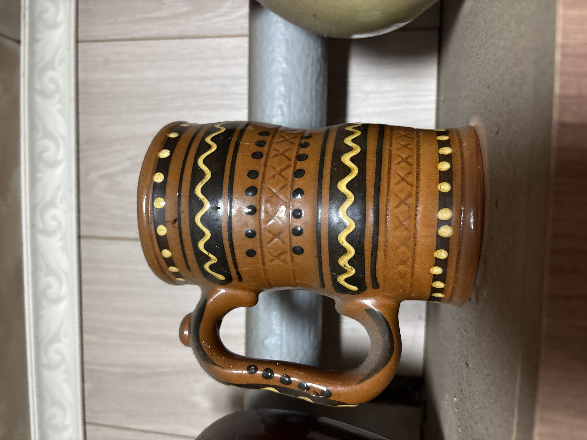 The image shows a close-up, eye-level, full shot of a brown ceramic mug with black and yellow decorative patterns. The mug has a handle and appears to be sitting on a shelf. The background consists of vertical wooden planks and a decorative white trim at the top. To the left of the mug, a dark, indistinct object is partially visible, and to the right, a pale green spherical object is visible. The shelf itself appears to be made of a textured material. The lighting is even, suggesting an indoor environment. There are no people or animals present in the image. The location is specified as Ветлужский, Russia.