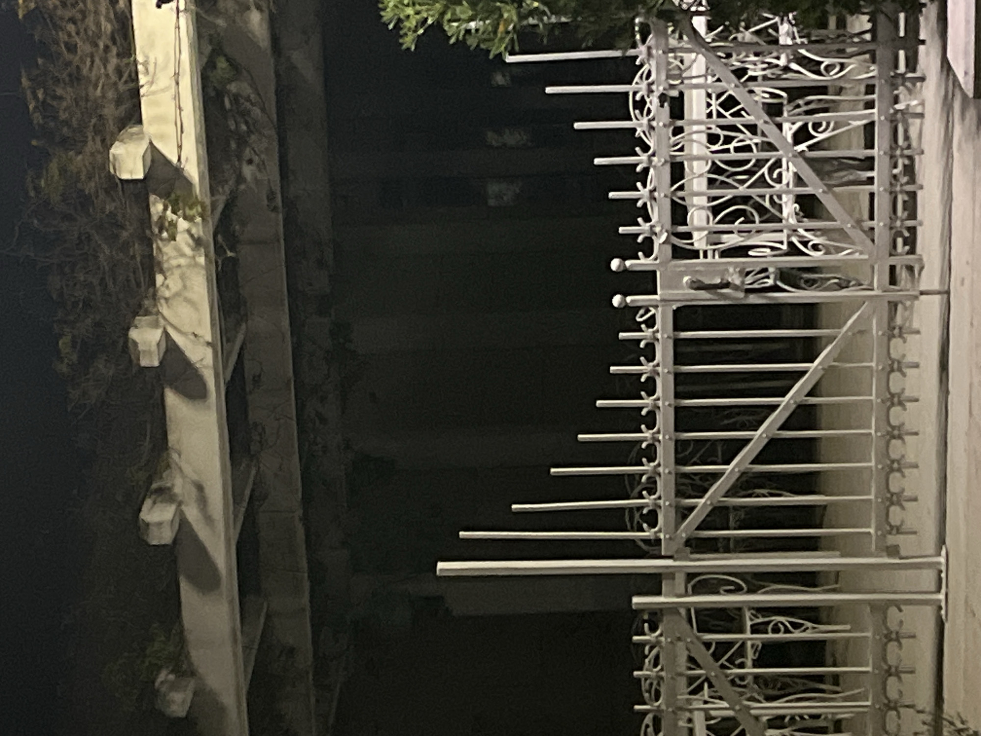 An eye-level, indoor photograph depicts a wrought iron balcony against a dark background. The balcony features intricate designs, including horizontal bars with decorative elements and diagonal supports. To the left, a building facade with a white, decorative vertical element is visible. The scene is dimly lit, suggesting it is nighttime. The location is Budva, Montenegro.
