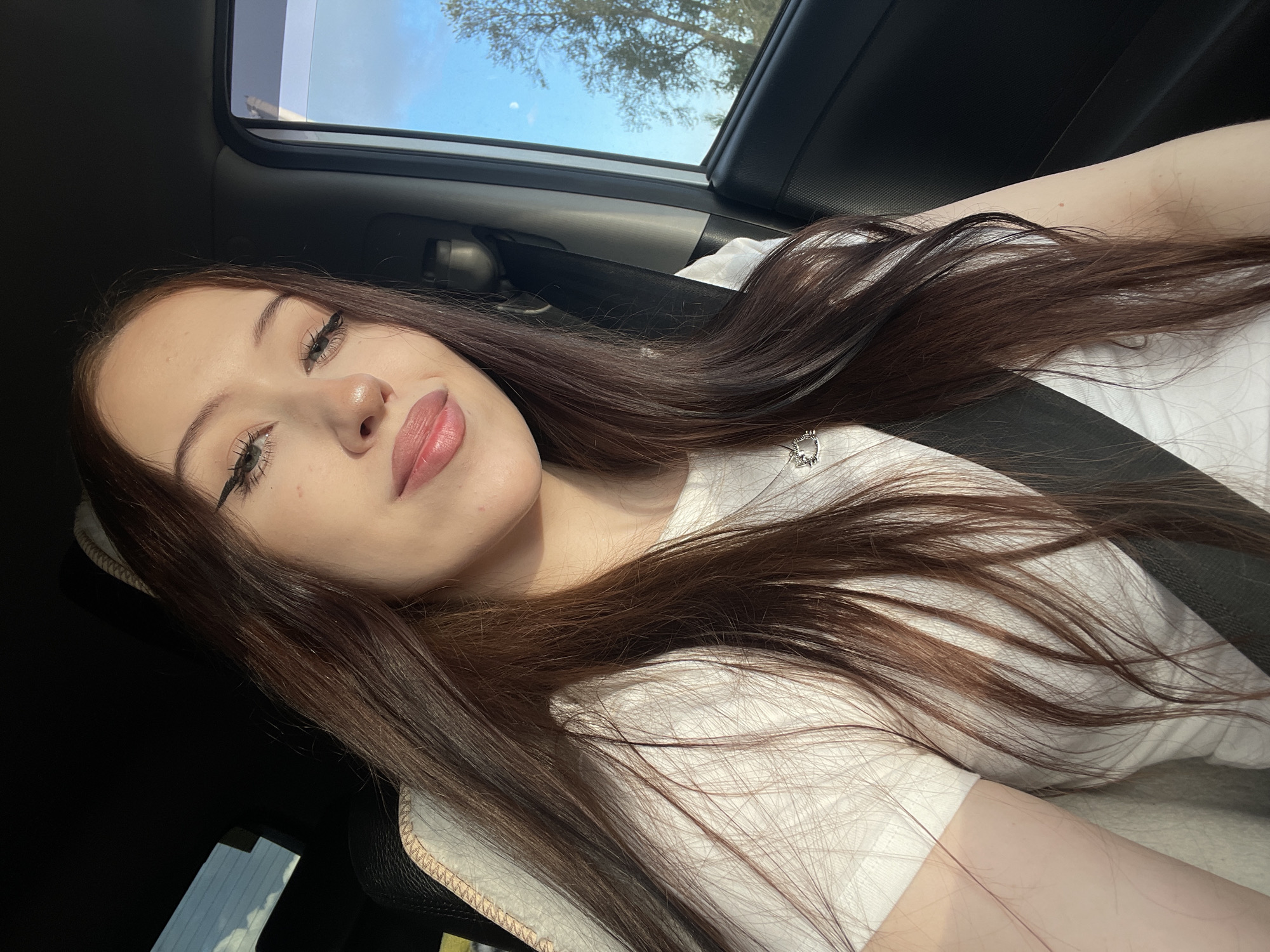 A young woman with long, dark brown hair and fair skin is seen from a selfie perspective inside a car. She is wearing a white t-shirt and has subtle makeup, including winged eyeliner. Her hair is spread across her chest and shoulder. She appears to be looking at the camera with a slight smile.
The scene is illuminated by natural light, suggesting it is daytime. Sunlight is coming from the right, casting shadows and highlighting her features. Through the car's sunroof and possibly window, a bright blue sky with a few wispy clouds is visible, along with the silhouette of tree branches, indicating an outdoor setting. The car's interior is dimly lit in comparison, with a black dashboard and a grey seatbelt partially visible. A beige-colored edge of the car seat is also in the foreground. The overall mood appears relaxed and casual.
There is no visible text in the image.
