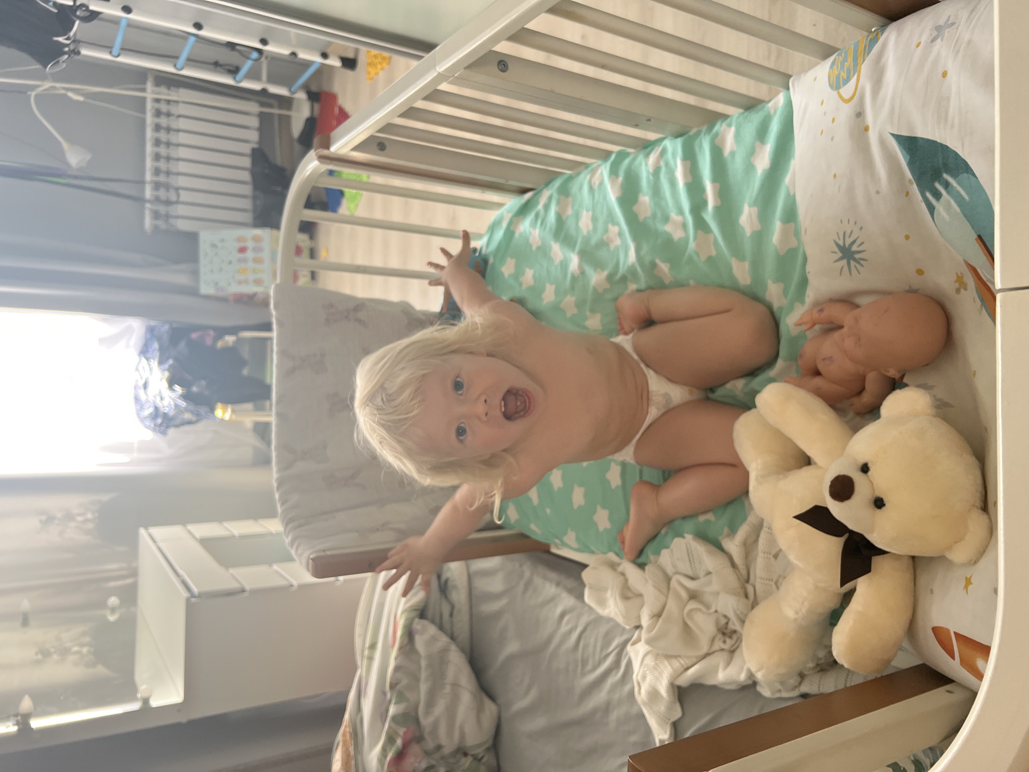 This image, rotated 90 degrees counter-clockwise, features a lively blonde-haired toddler lying on their back inside a white wooden crib. The child, wearing only a white diaper, looks directly at the camera with wide blue eyes and an open mouth, appearing to be in mid-shout or laugh, with arms playfully outstretched.
The crib is set against a brightly lit backdrop, likely a window to the left, indicating daytime in Sochi, Russia. Inside the crib, the toddler is surrounded by a mint green fitted sheet adorned with white star patterns and a white comforter featuring space-themed illustrations like planets and a rocket ship. Two toys accompany the child: a small, light-skinned baby doll near the child's head and a larger, cream-colored teddy bear with a dark brown bow around its neck, resting near the child's feet. A crumpled white knitted item is also visible within the crib.
In the slightly blurred background, elements of a domestic setting can be seen, including a white clothes drying rack and other pieces of light-colored furniture. The scene captures a candid and joyful moment, highlighting the child's active and expressive personality within a comfortable and child-friendly environment. There is no visible text in the image.