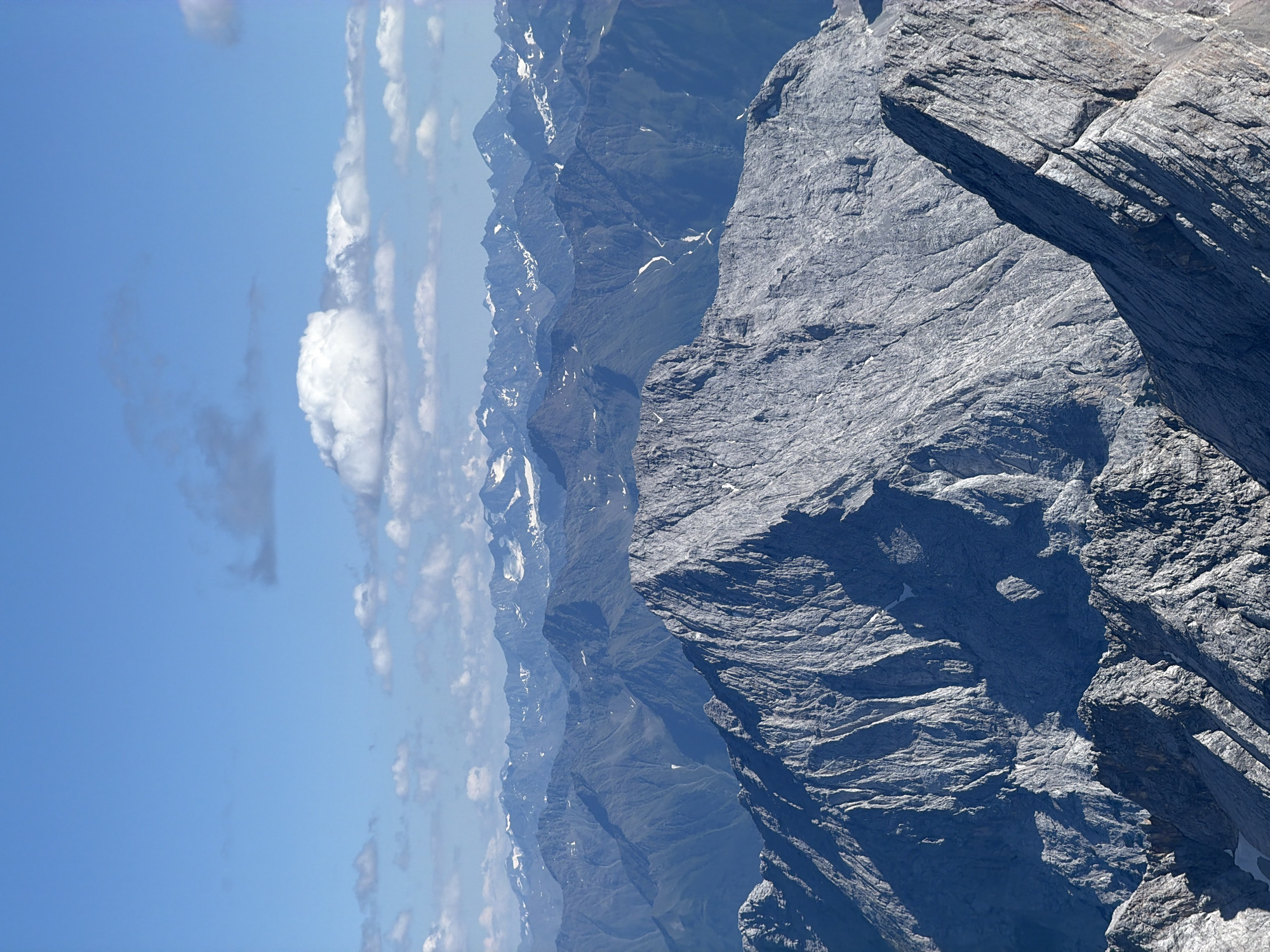 The image depicts a vast mountain range under a bright blue sky with scattered white clouds. In the foreground, a jagged, rocky mountain peak dominates the lower portion of the frame, casting sharp shadows. Behind it, layers of progressively more distant mountain ranges recede into the horizon, their surfaces textured with slopes, ravines, and patches of snow, particularly on the higher peaks. The overall impression is one of immense scale and rugged natural beauty. The image appears to be taken from a high vantage point, looking out over the mountainous terrain surrounding Grainau, Germany, likely a view of the Alps. There are no people or man-made objects visible in the scene.