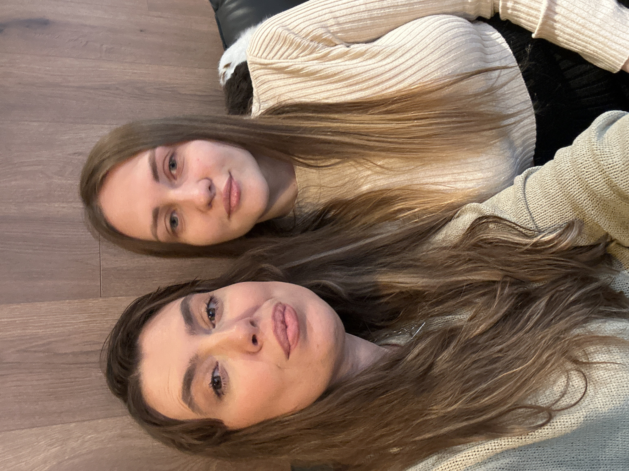Two women lie side by side on a wooden floor. The woman on the left has dark brown hair, while the woman on the right has light brown hair. The woman on the left is wearing a light-colored sweater. The woman on the right is wearing a beige, ribbed sweater. A portion of a black, likely leather, object is visible behind the woman on the right. The location cannot be verified in Batumi, Georgia based on the information provided.