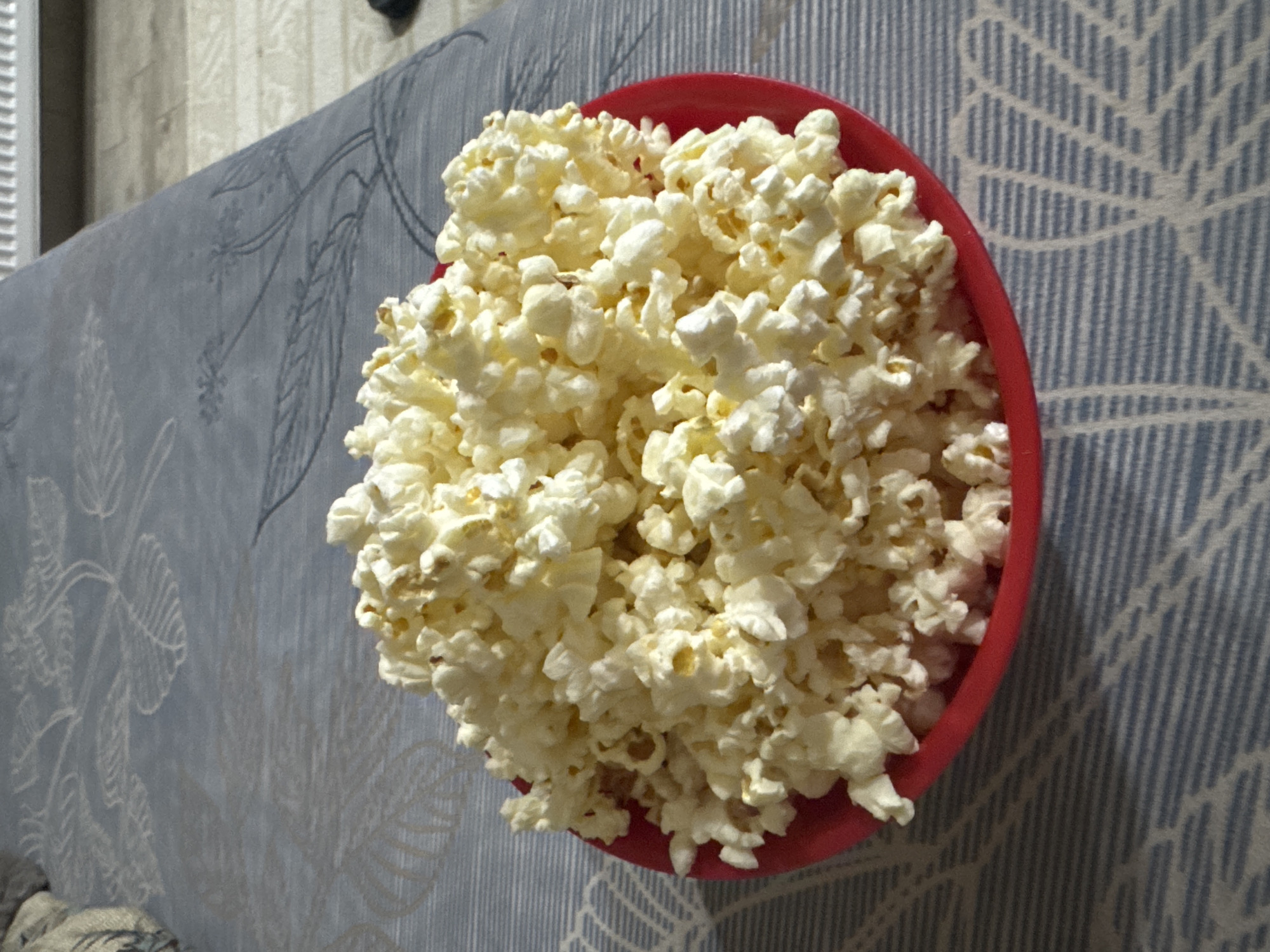 This media file presents an overhead view of a red bowl brimming with freshly popped, white popcorn. The popcorn fills the bowl to capacity, creating a mounded peak. The bowl is placed on a fabric surface, likely upholstery or a blanket, which serves as the background. This fabric features a distinct pattern of light blue and white vertical stripes, subtly adorned with lighter, stylized leaf or botanical designs. The lighting in the image appears to be artificial and internal, suggesting an indoor setting without strong natural light. There are no people visible, and no specific action is taking place beyond the presence of the popcorn, which typically implies a context of casual relaxation or entertainment, such as watching a movie. No visible text is present in the image. Although the context provided indicates the location is Lehchevo, Bulgaria, there are no visual elements within the image itself to confirm this geographical detail.