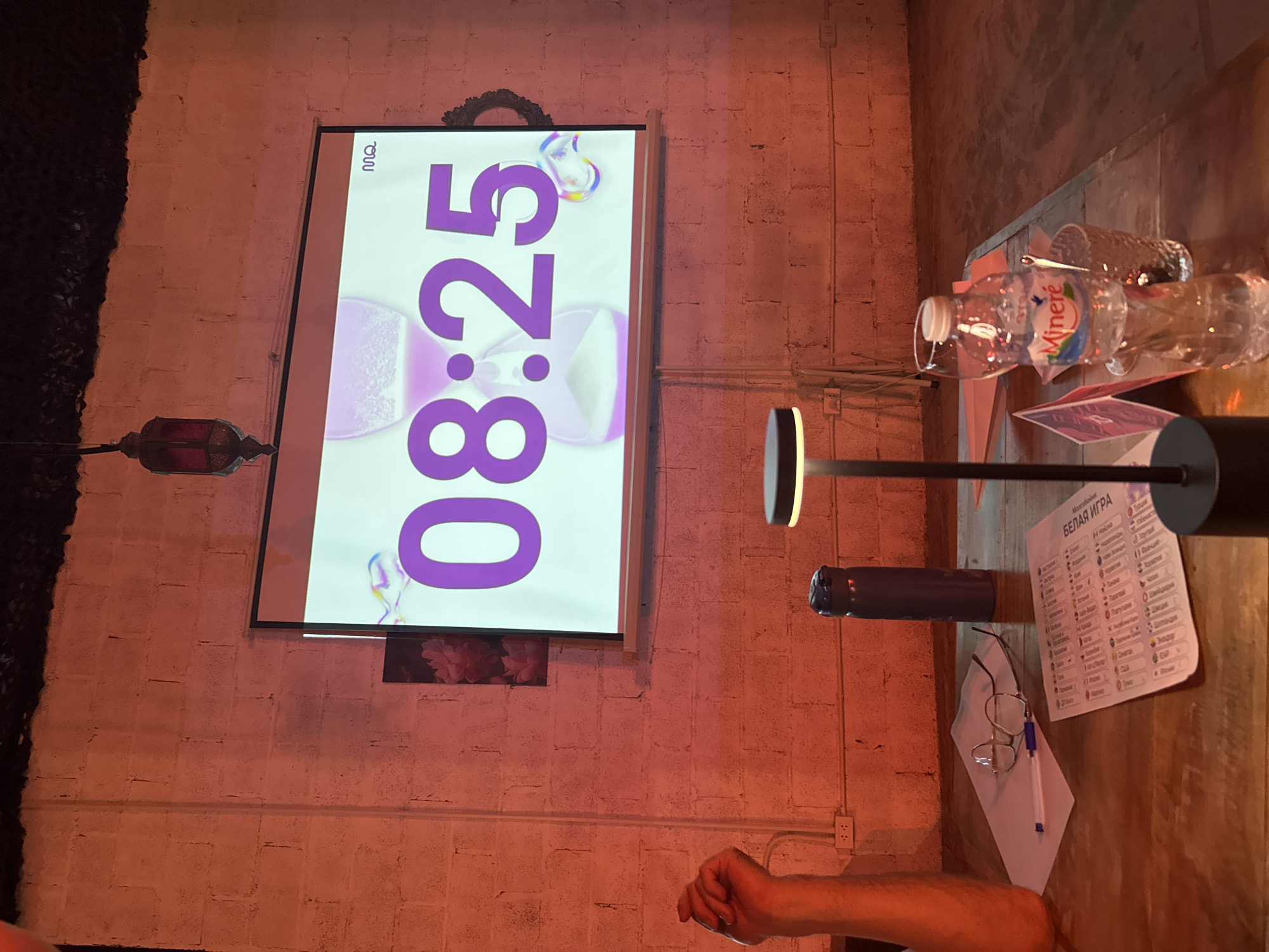 An overhead shot captures a rustic wooden table with various items, including a metallic thermos, a pair of glasses, a pen, and a paper document. A bottle of Minere water and a glass of crushed ice are also present. A projection screen displays a purple hourglass graphic with the time "08:25" in large purple numerals. The background features a brick wall illuminated by warm lighting. A person's arm is visible on the left side of the frame, with their fist raised. A desk lamp with a modern design stands on the right side of the table. The overall setting suggests an informal gathering or event.