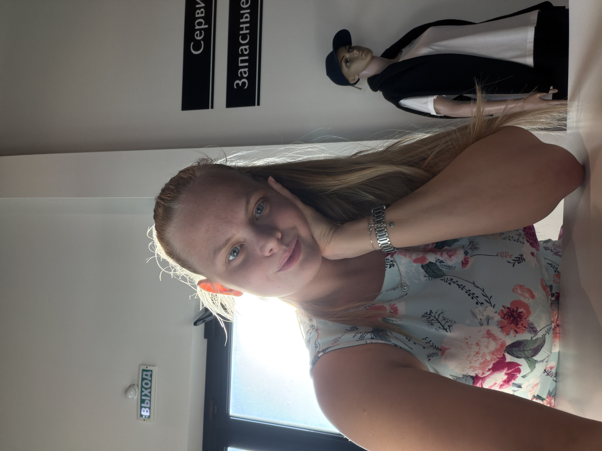 A blonde-haired woman takes a selfie in a building in Izhevsk, Russia. She is wearing a floral dress and a watch. In the background, there is a mannequin wearing a black hat and a black and white coat. There are signs on the wall in Russian that read "Сервис" and "Запасные". There is also an exit sign in Russian. The woman is touching her face with her hand.